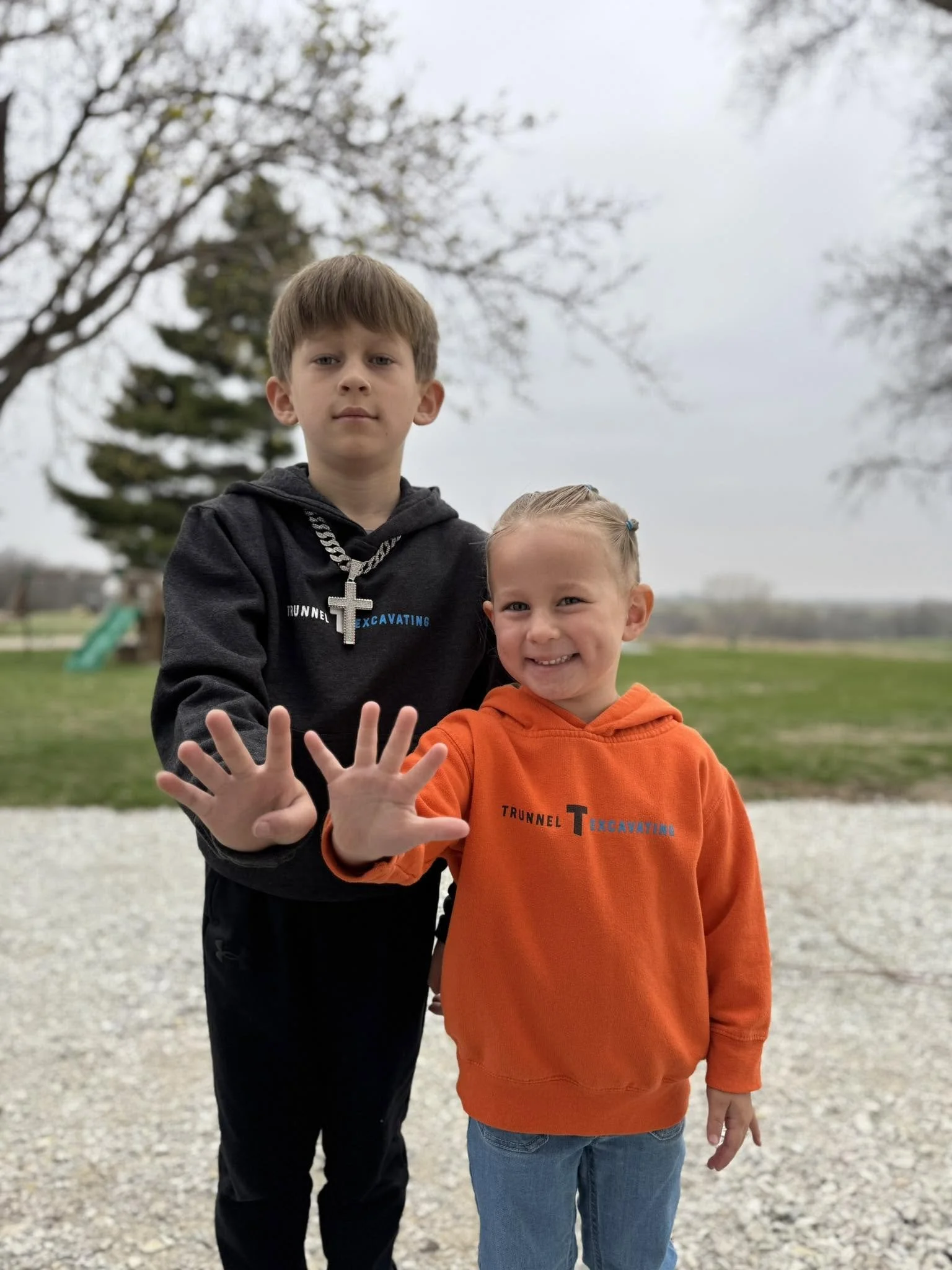 Two kids, one taller with brown hair and a cross necklace, wearing a black hoodie, and a smaller with blonde hair and an orange hoodie, smiling and holding up their hands in a playful gesture outdoors on a cloudy day with trees in the background.