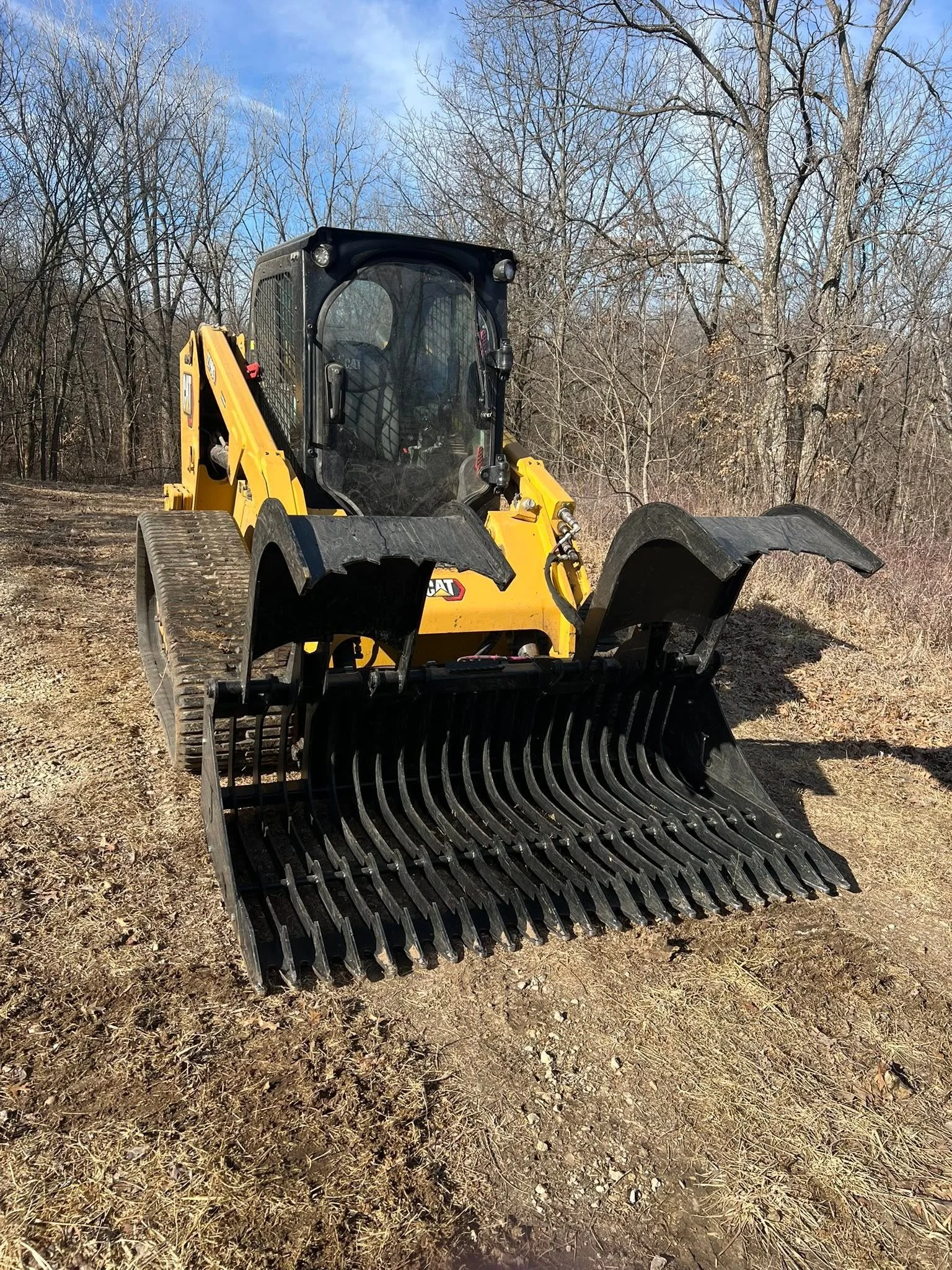 A yellow Bobcat skid-steer loader with a black protective cab, parked on a dirt patch with leafless trees and a cloudy sky in the background.