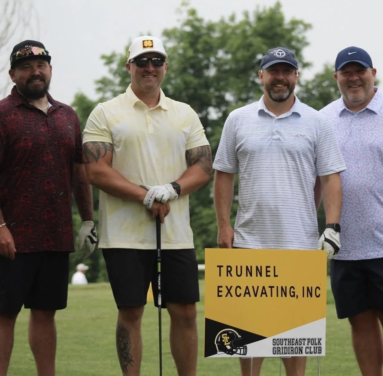 Four men standing outdoors on a golf course, smiling, next to a yellow sign that reads "Trunnel Excavating, Inc." and "Southeast Polk Gaidron Club," with trees in the background.