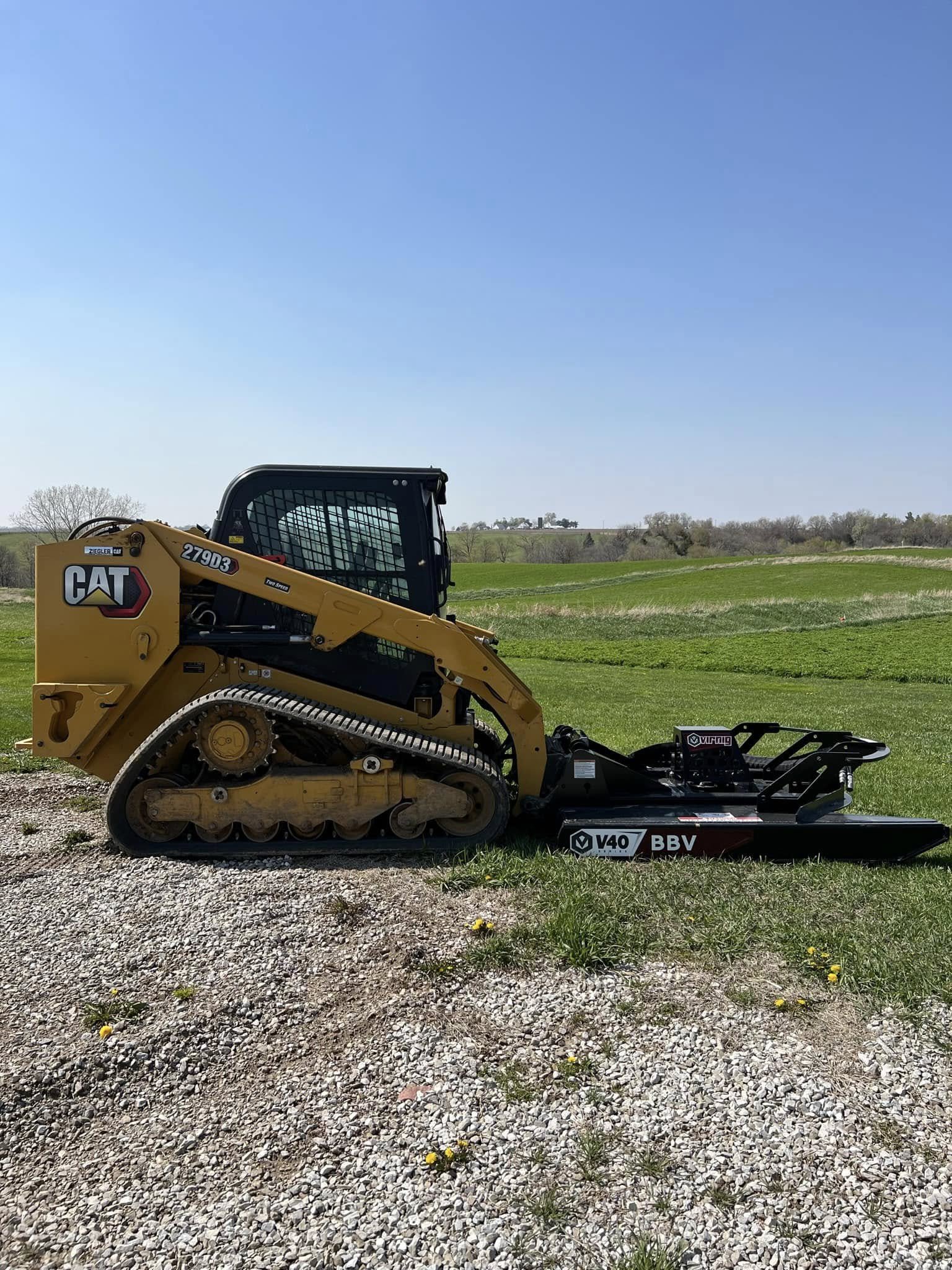 A yellow Caterpillar compact skid steer loader on a gravel surface in a rural field with green grass, trees, and a blue sky in the background.