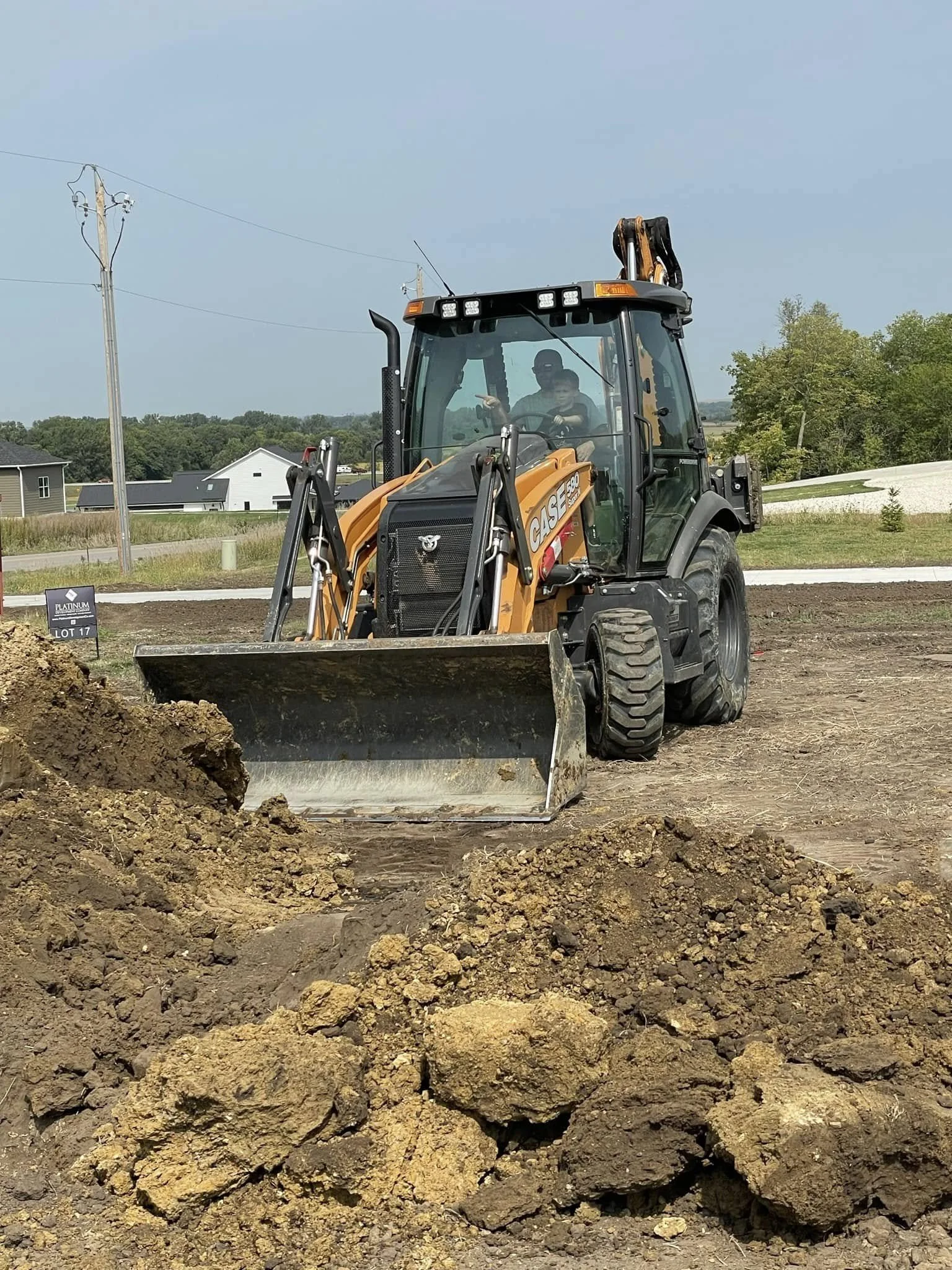 A construction site with a backhoe loader digging into the soil, with two workers inside the cab. There are houses and trees in the background under a cloudy sky.