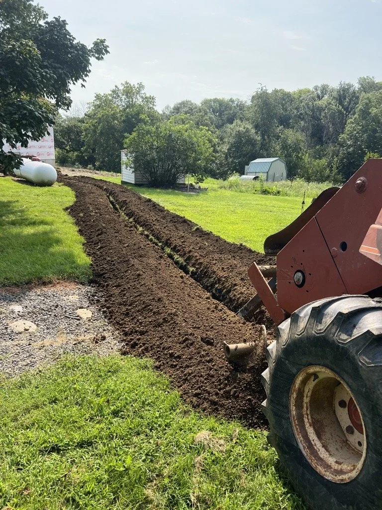 A construction vehicle is digging a trench in a grassy field with small trees and sheds in the background.