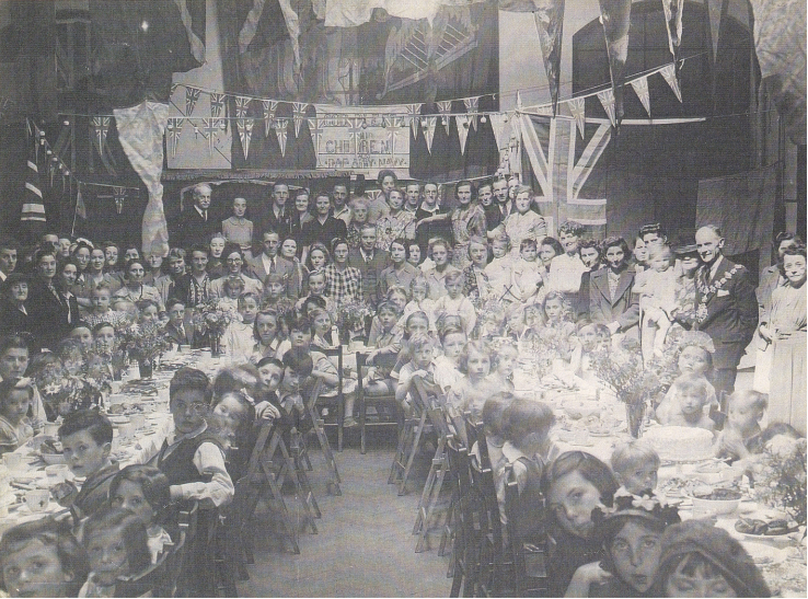 Greenwich Central School hall: A large group of adults and children gather around long tables in Greenwich Central School hall, decorated with bunting and Union flags. The Mayor of Greenwich and the landlady of the Royal Albert pub are among the grou