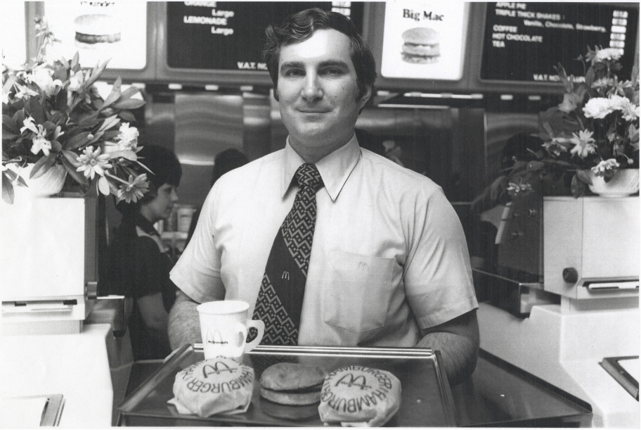 McDonald’s Woolwich, 13 November 1974: Man in a shirt and McDonald’s-logo tie holds a tray with wrapped burgers, a drink and a Big Mac in the first UK McDonald’s branch in Woolwich. Menu boards and floral displays are visible behind him.
