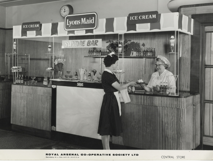 Lyons Maid Sundae Bar, 1963: Woman customer at a Lyons Maid ice-cream counter is served by a staff member in a white uniform. Bottles, sundae glasses and ice-cream displays line the counter beneath a “Lyons Maid Ice Cream” sign.