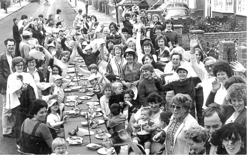 Bracondale Road street party, 1981: Residents of Bracondale Road, Abbey Wood, sit and stand around a long outdoor table for a street party celebrating the 1981 royal wedding, waving towards the camera.