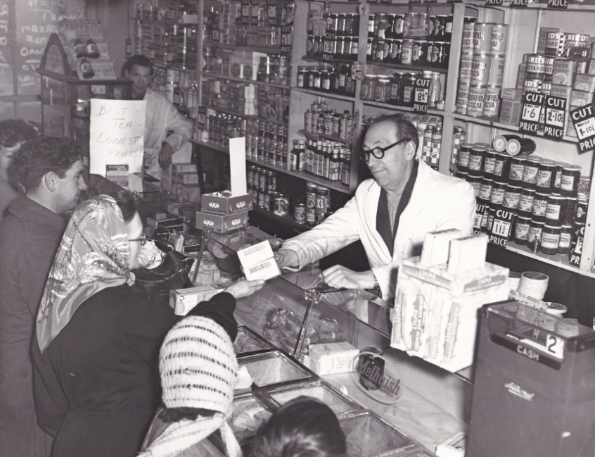 Alfie Bass’s grocery shop: Alfie Bass serves customers from behind the counter of his grocery shop on Greenwich Church Street. The small shop is packed with jars, tins, tobacco products and handwritten price signs.