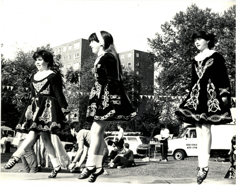Irish dancers, Charlton House, 1980. 
Three dancers in embroidered traditional Irish dresses perform outdoors at Charlton House, with spectators, bunting and residential buildings in the background.
