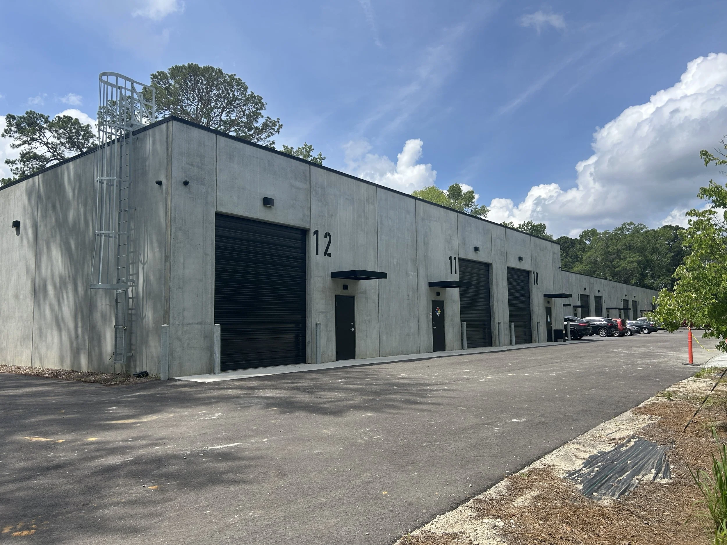 A row of large industrial storage units with numbered roll-up doors, numbered 10, 11, and 12, outside in a parking lot with cars, surrounded by trees and a blue sky with clouds.