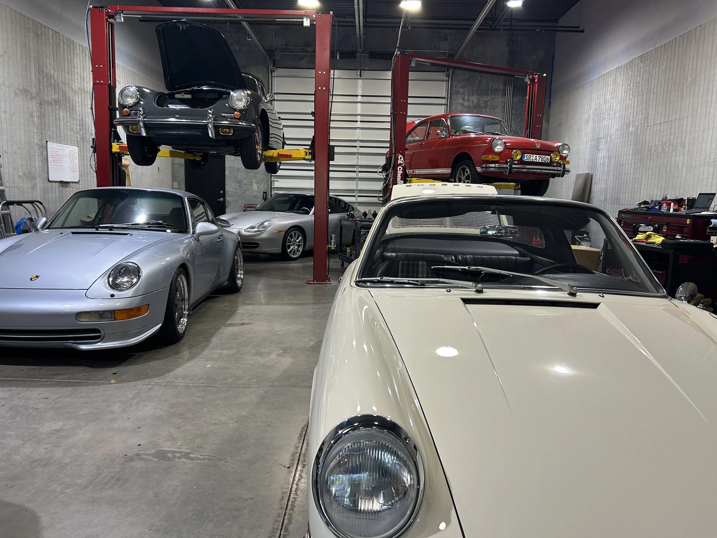 Multiple cars inside a garage with some cars on lifts, including a gray vintage Porsche and a red classic Volkswagen Karmann Ghia.