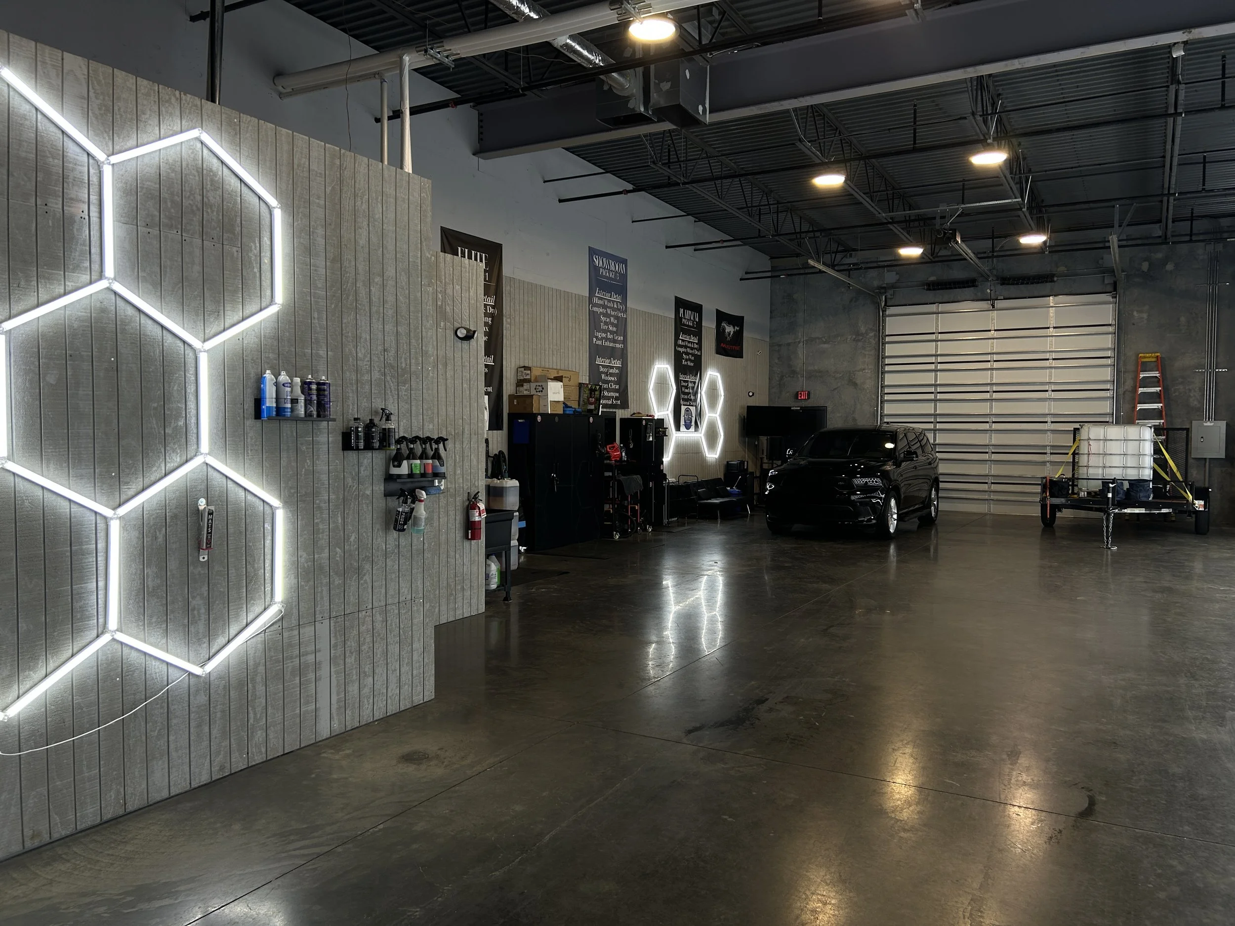 Interior of a modern industrial-style garage or workshop with concrete floors, metal walls, and a sports car parked inside. Neon light fixtures form hexagon shapes on the wall, and there is a large garage door at the back. Various tools and equipment