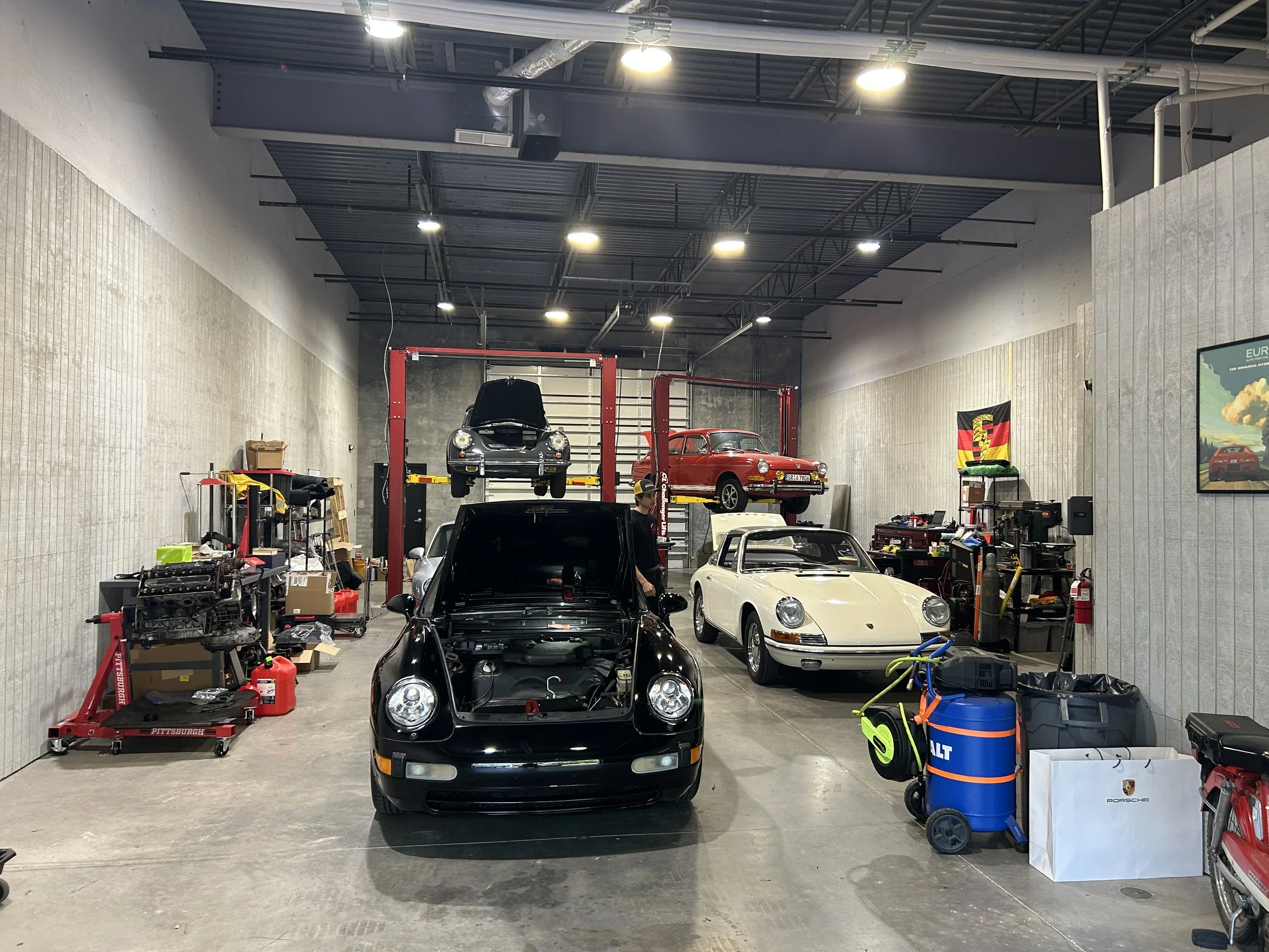Indoor car workshop with several Porsche cars being serviced, including one on a lift with its hood open and others parked inside, surrounded by tools and equipment.