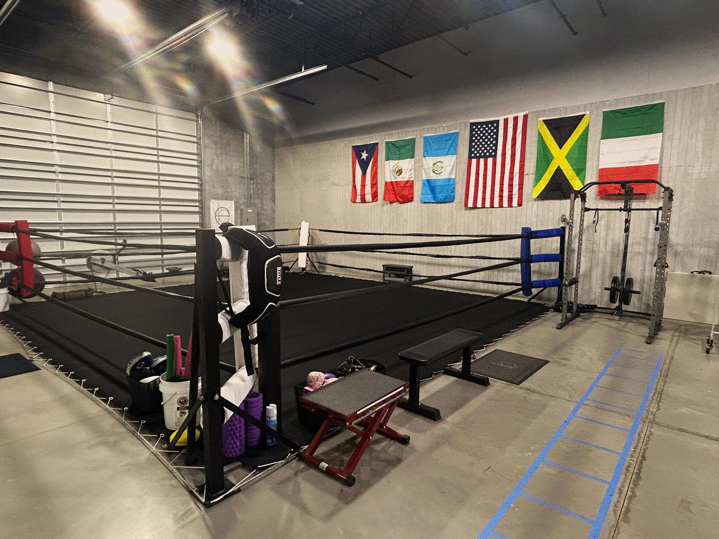 Inside a boxing gym with a boxing ring, training equipment, and flags hanging on the wall, including flags of Puerto Rico, Mexico, Guatemala, the United States, Jamaica, and Italy.
