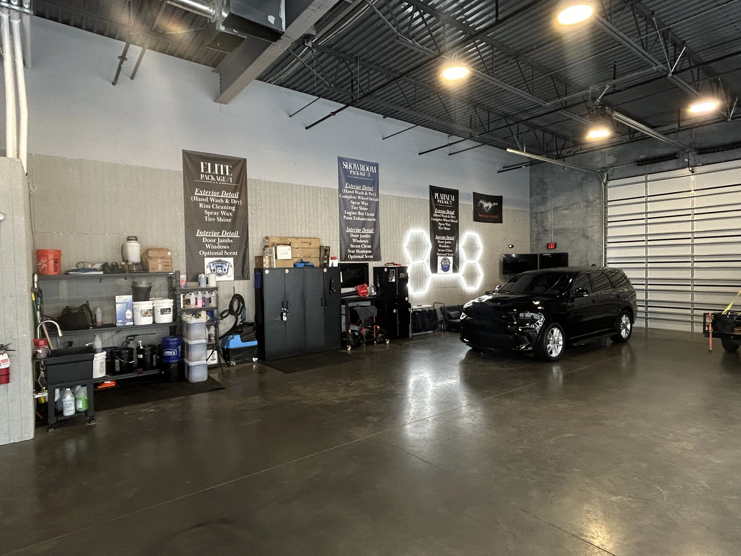 Inside a car detailing or auto repair shop with a black car parked on the right side and various tools and supplies on shelves on the left side. There are multiple promotional banners listing services on the wall above the shelves. The shop has a hig