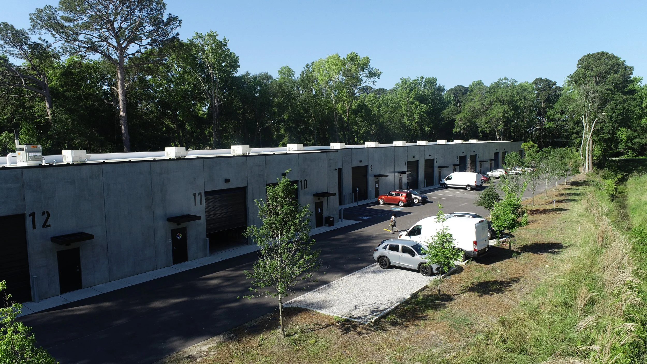 A row of storage units with numbered doors in a parking lot surrounded by green trees and grass, with several cars parked and a person walking.