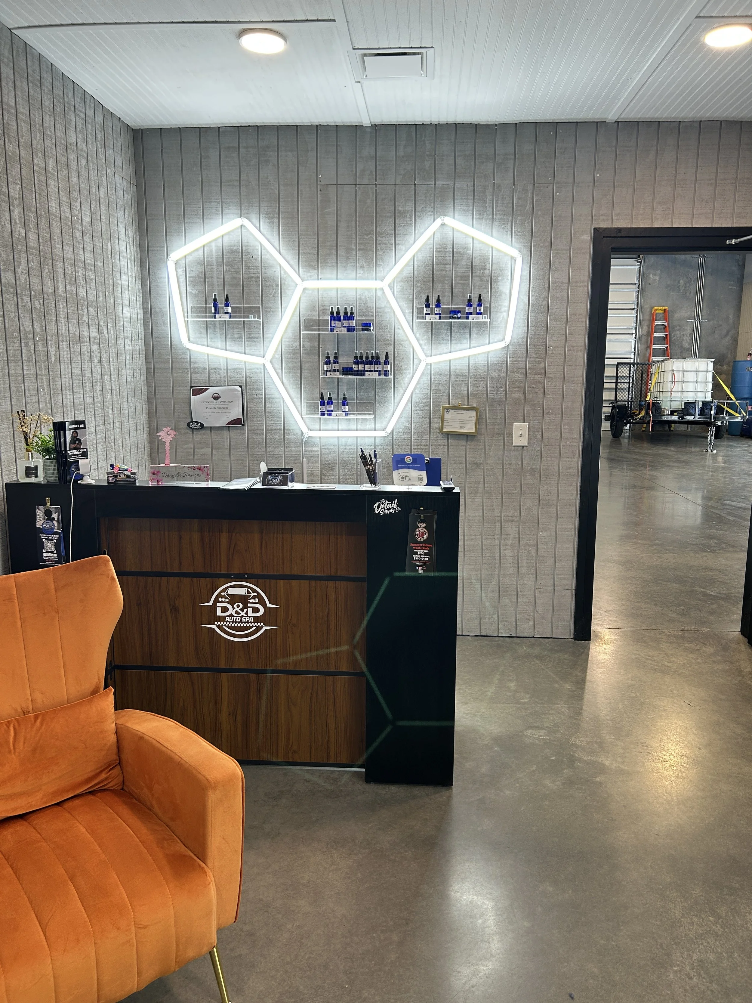 A waiting area with an orange sofa, a reception desk with a logo reading 'D&D Auto Spa,' and a decorative wall behind the desk with a neon light design resembling interconnected hexagons and bottles on shelves.