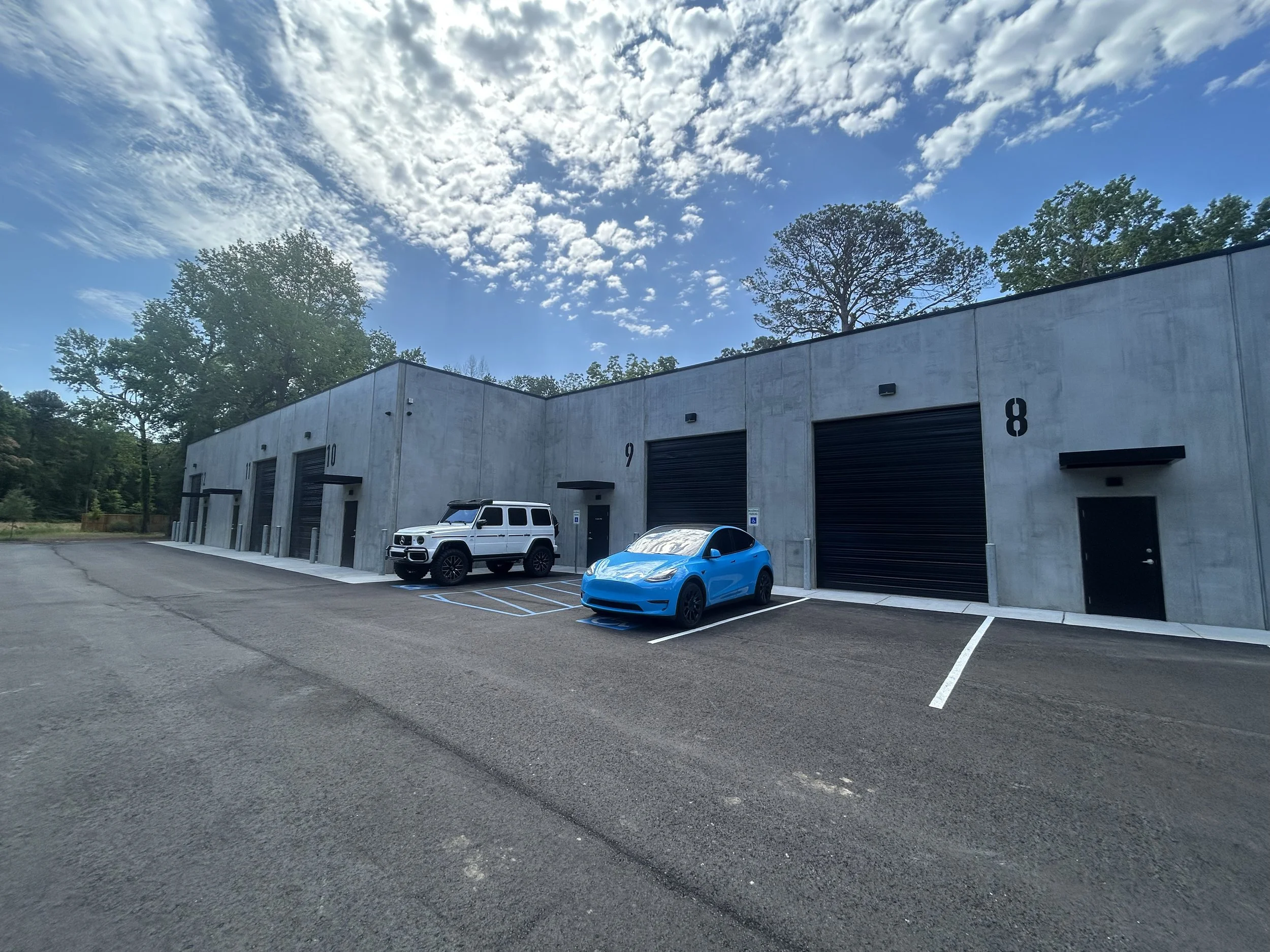 A row of storage units with black roll-up doors, parking spaces in front, and a white SUV and a blue car parked, set against a backdrop of trees and a partly cloudy sky.