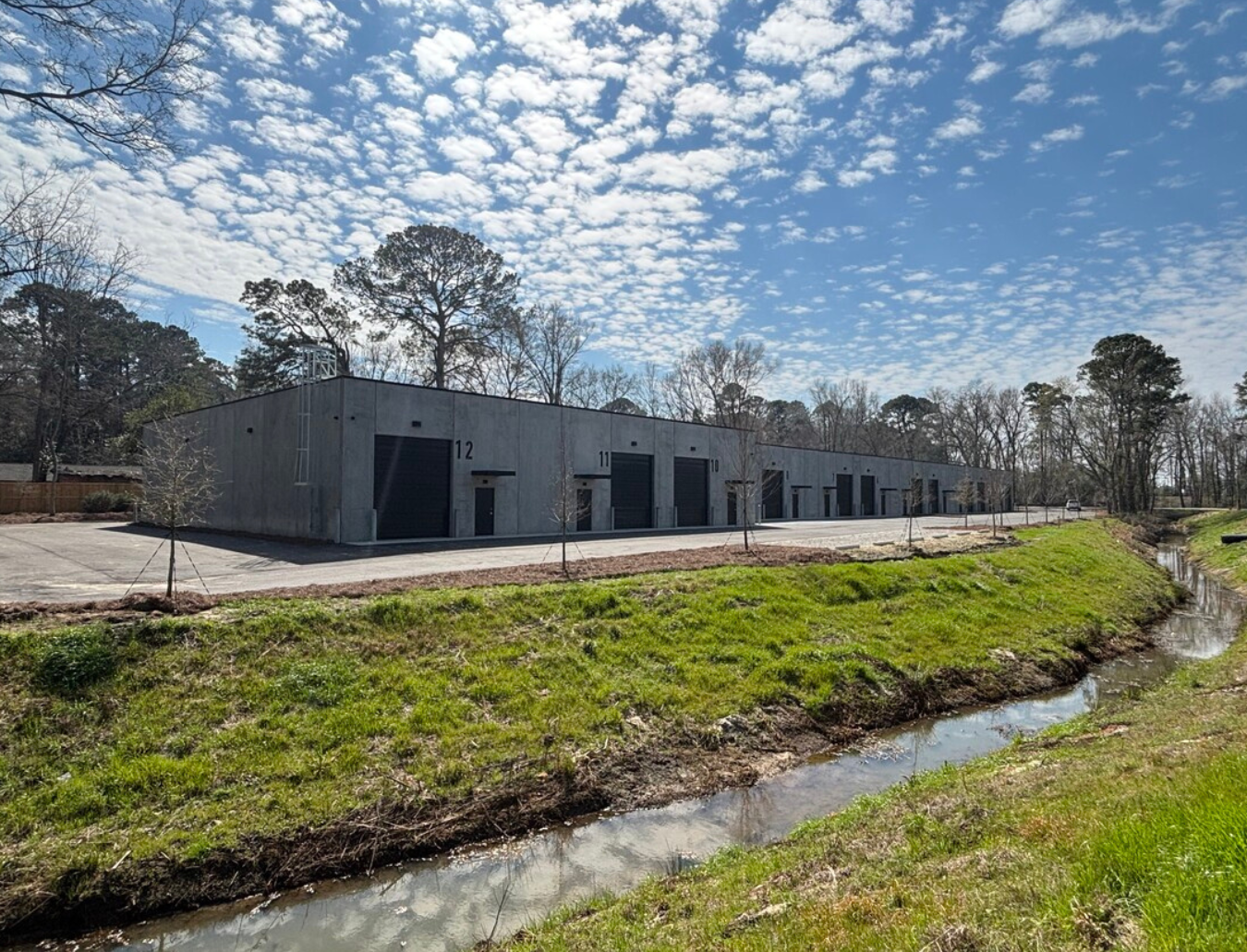 A modern warehouse building with numbered doors, surrounded by young trees, next to a small creek, under partly cloudy sky.