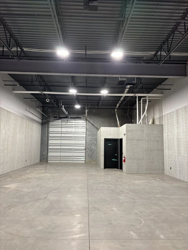 Empty industrial space with concrete floor, high ceiling, overhead lights, a large garage door, and a small enclosed room with a black door and fire extinguisher.