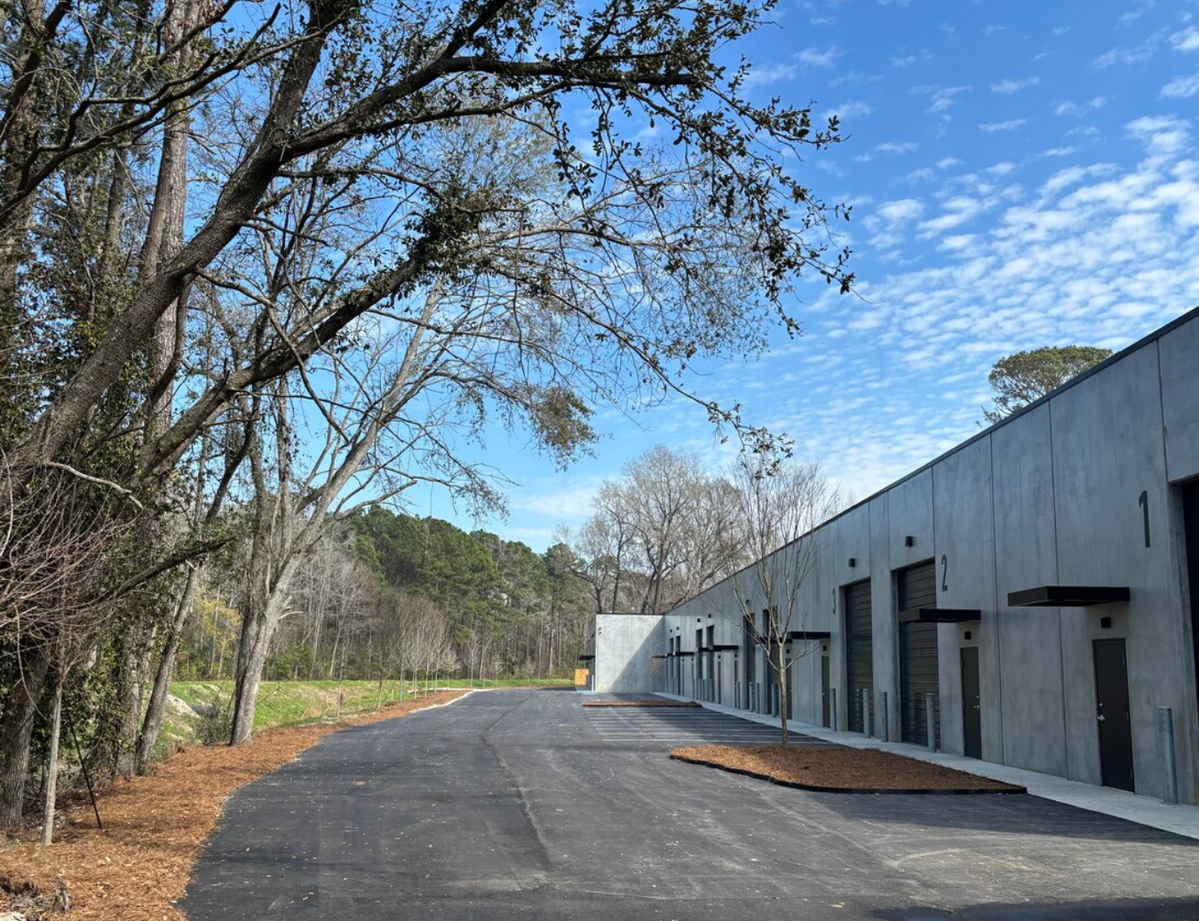 Industrial warehouse with numbered loading dock doors, surrounded by young trees, on a clear day with blue sky and scattered clouds.