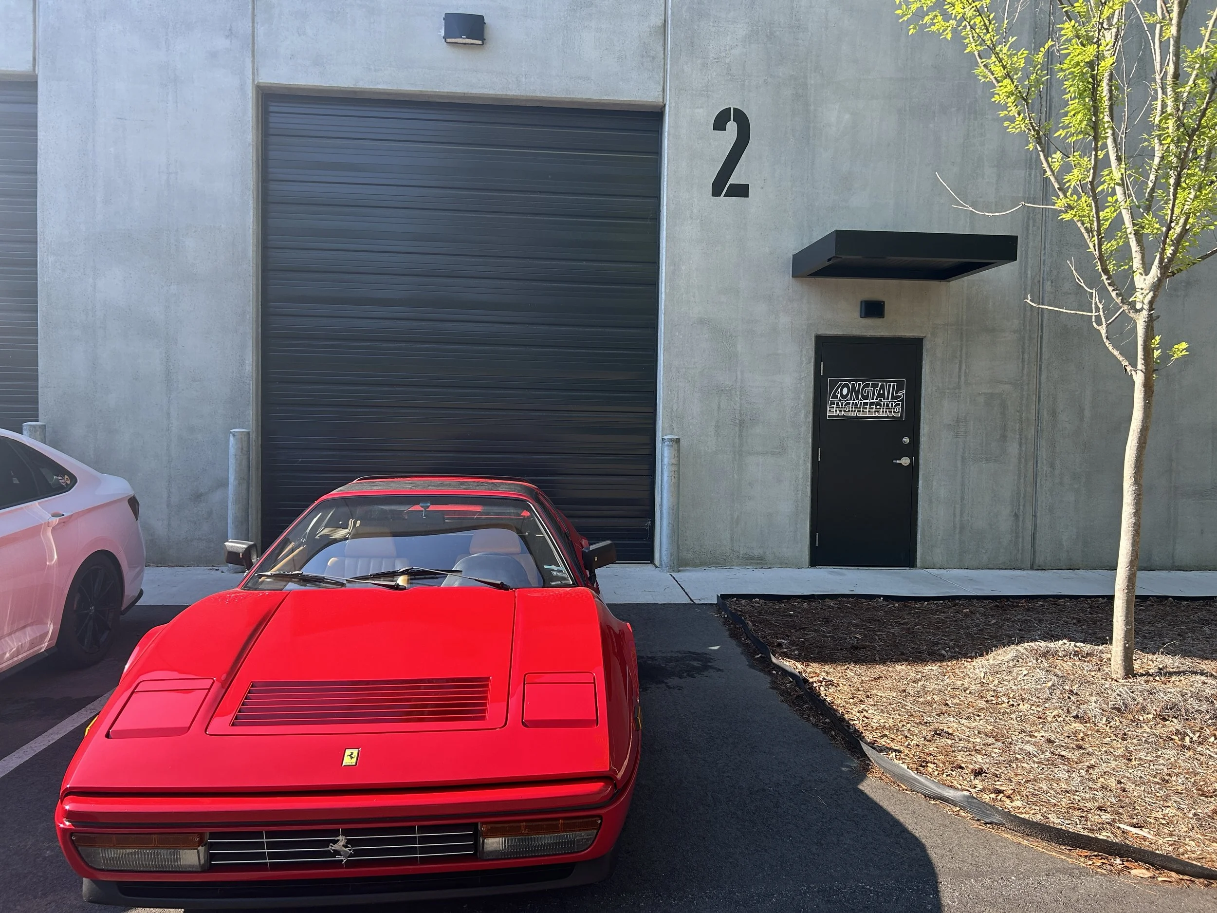 A red Ferrari sports car parked in front of a modern building with a gray concrete facade. The building has a black garage door, a side door with the words 'Longtail Engineering,' a large black number '2' on the wall, and a small overhang above the s