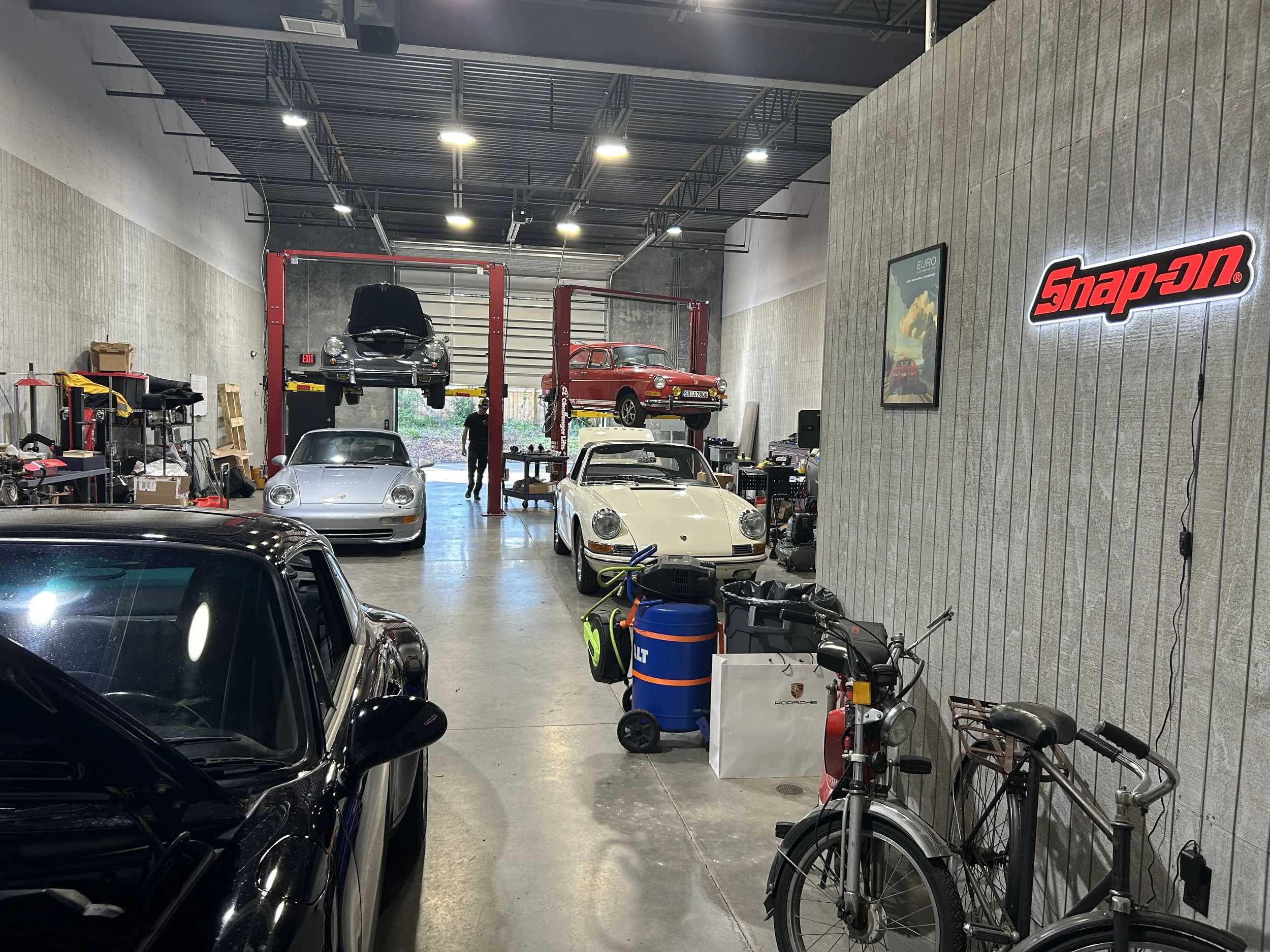 Car workshop with several cars, including a black Porsche with open hood, a silver Porsche, and a red classic car on a lift. Bicycles, tools, and equipment are scattered around, with a large Snap-on sign on the wall.