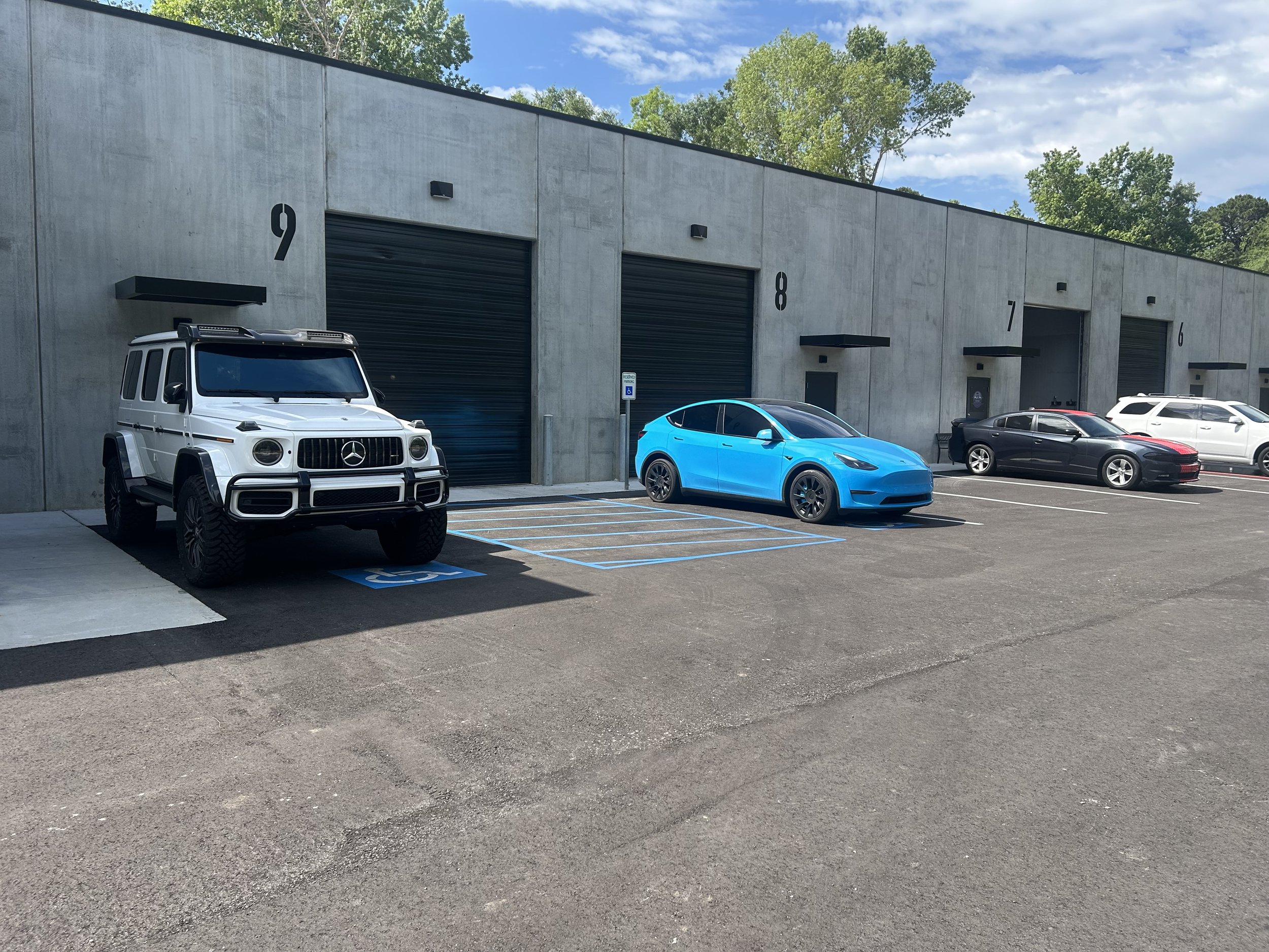A parking lot outside a concrete building with numbered garage doors, empty parking spaces marked with blue lines, and a few parked cars including a white Mercedes G-Class, a blue electric vehicle, and a black sedan. Trees and a blue sky with clouds 
