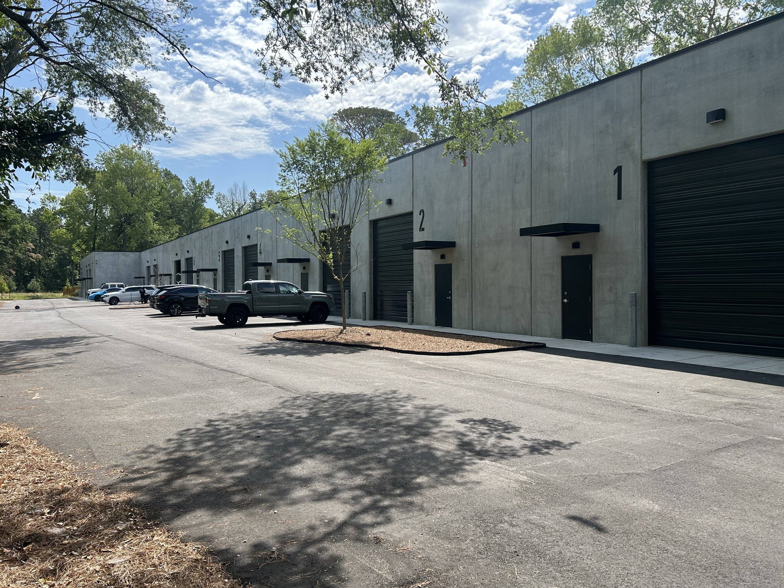 A row of industrial storage units with black roll-up doors, numbered 1 to 5, on a sunny day with a partly cloudy sky. In front, there are parked cars and a small tree.