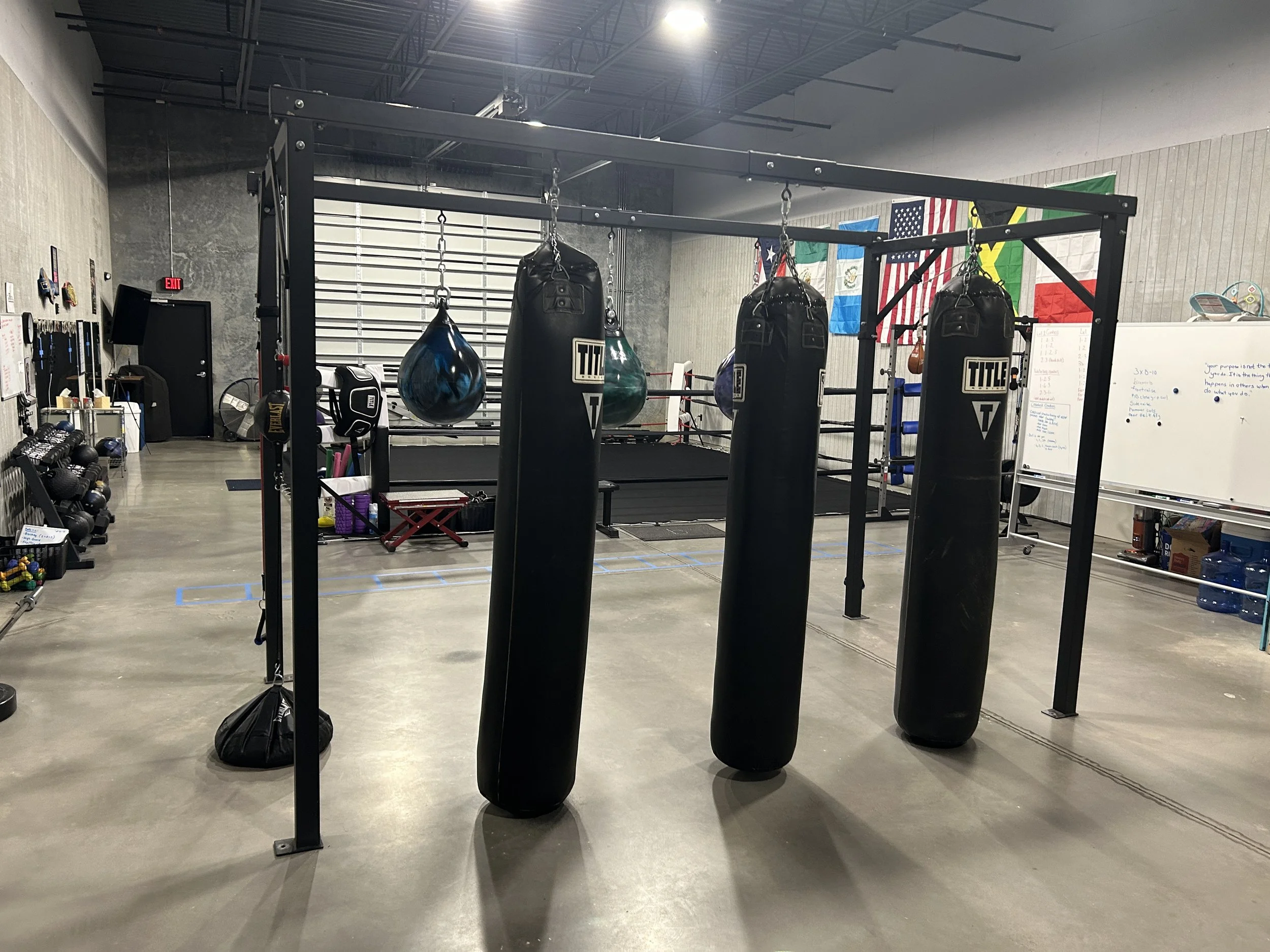 An empty boxing gym with several black punching bags hanging from a metal frame, American flags hanging on the wall, whiteboard with notes, and workout equipment on the side.
