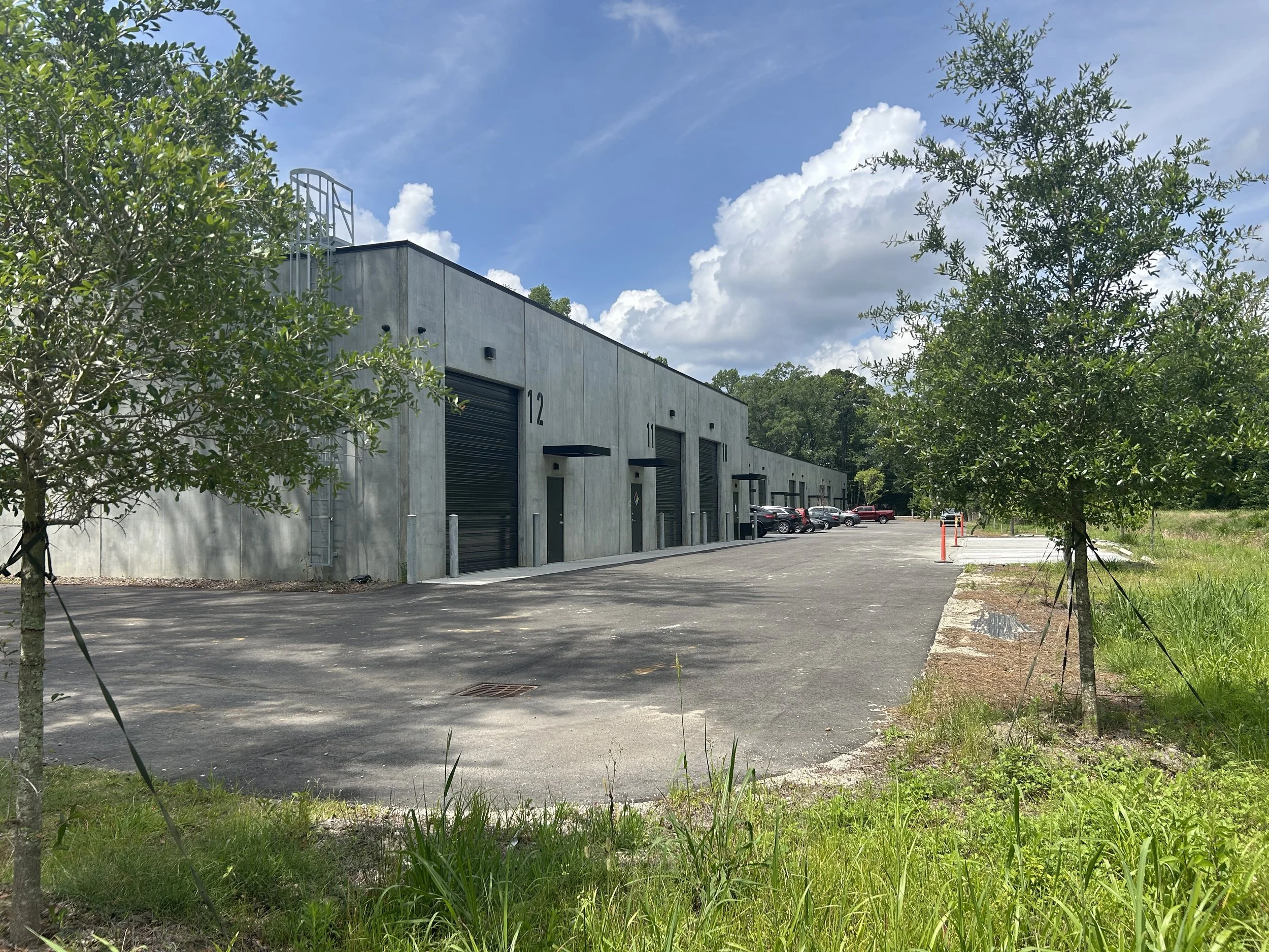 A concrete commercial building with numbered garage doors, parked cars, and surrounded by green trees and grass on a partly cloudy day.