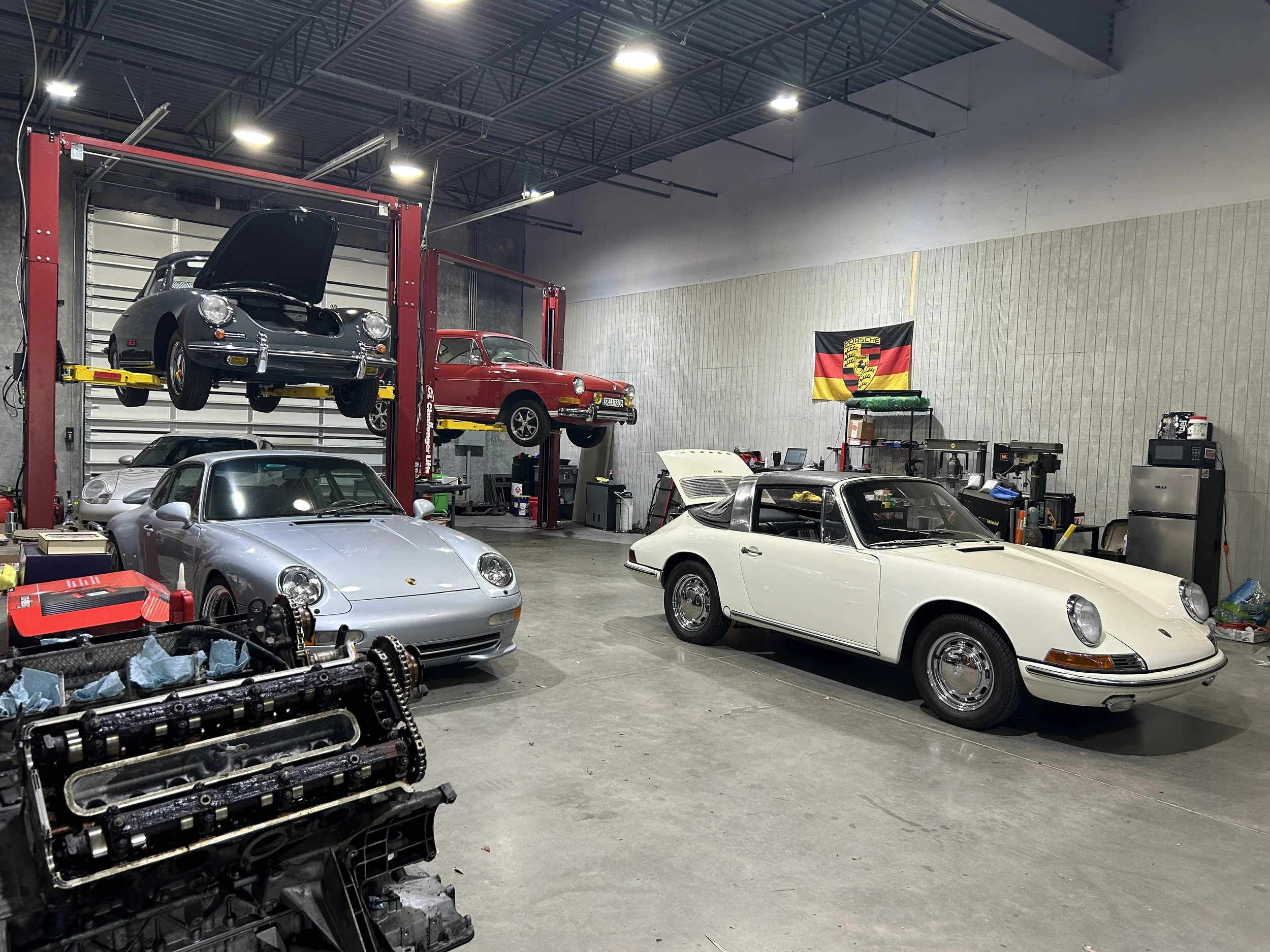 A garage workshop with multiple classic cars, including a silver Porsche and a red Porsche 911, some cars on lifts, and various tools and equipment around the space.