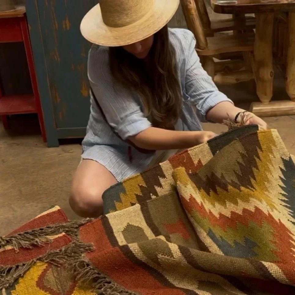 A person wearing a straw wide-brim hat and a striped shirt working on a colorful woven rug with geometric patterns in a rustic room.