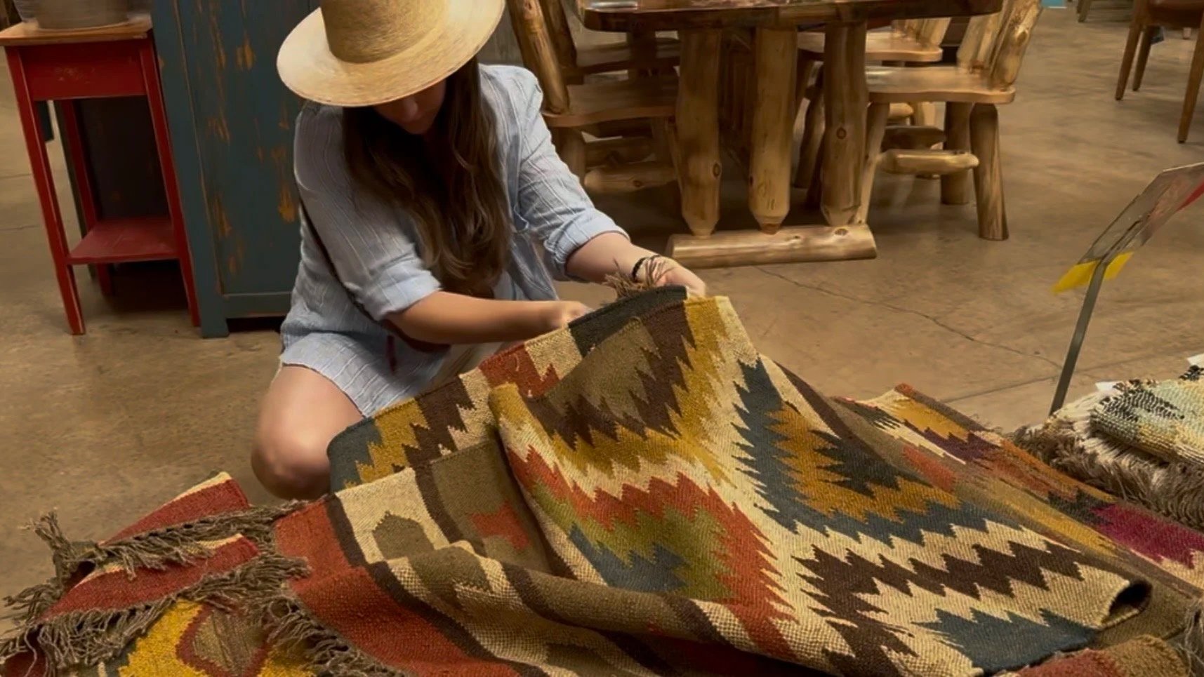 Woman sitting on the floor, wearing a hat, looking at a colorful woven rug in a store with wooden furniture.