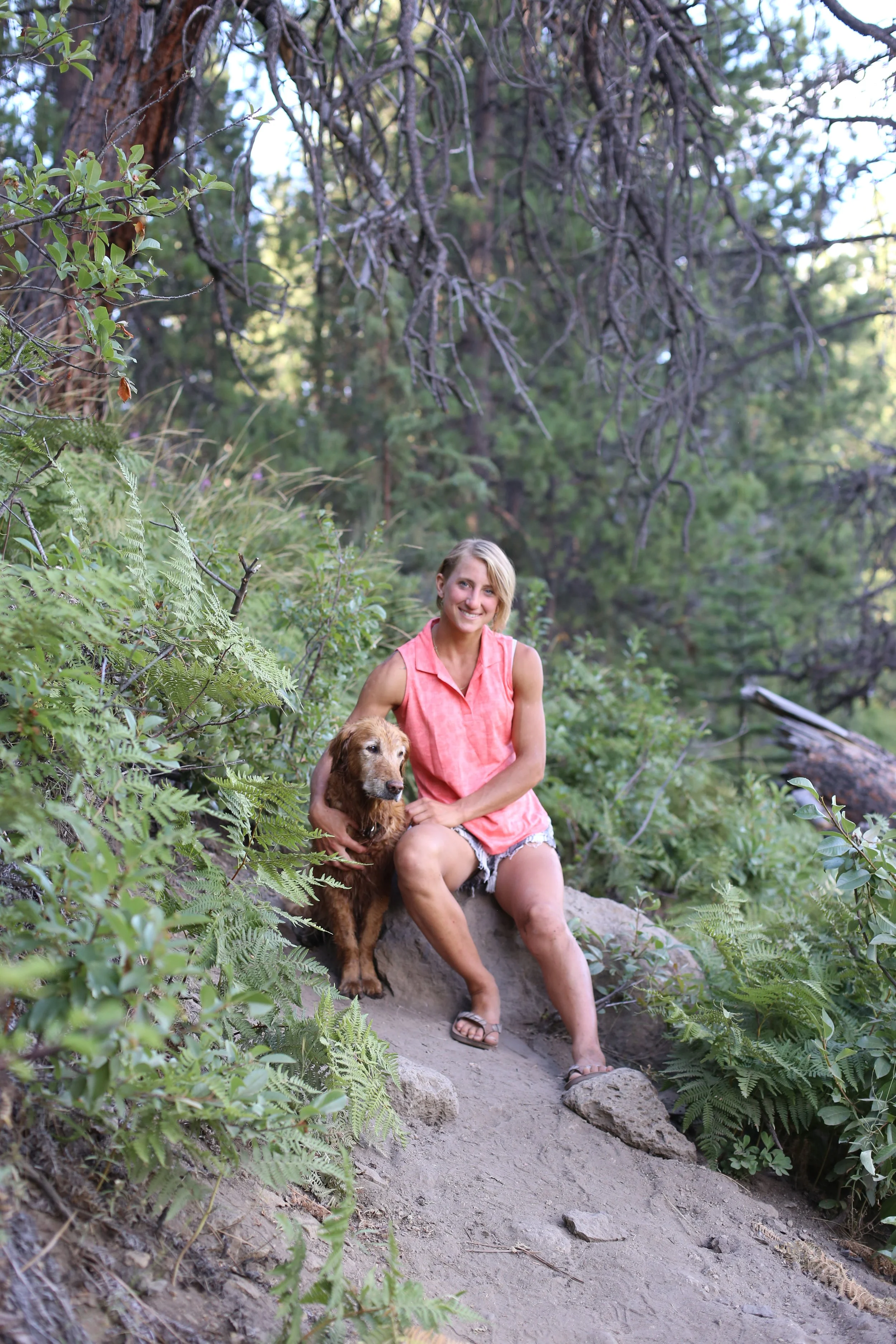 A smiling woman with short blonde hair, wearing a sleeveless coral top, shorts, and sandals, sitting on a rock in a forested area with green foliage and trees, holding a brown dog.