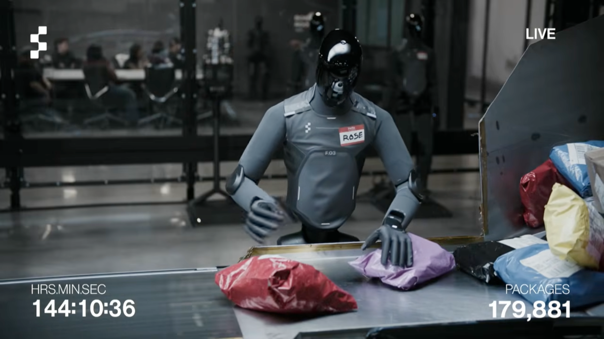 A humanoid robot with a black helmet and gray suit is sorting packages on a conveyor belt in a warehouse. There are several bags and boxes on the conveyor. In the background, people are seated at tables through a glass partition. The screen displays "LIVE" in the top right corner, along with numbers and a timestamp.