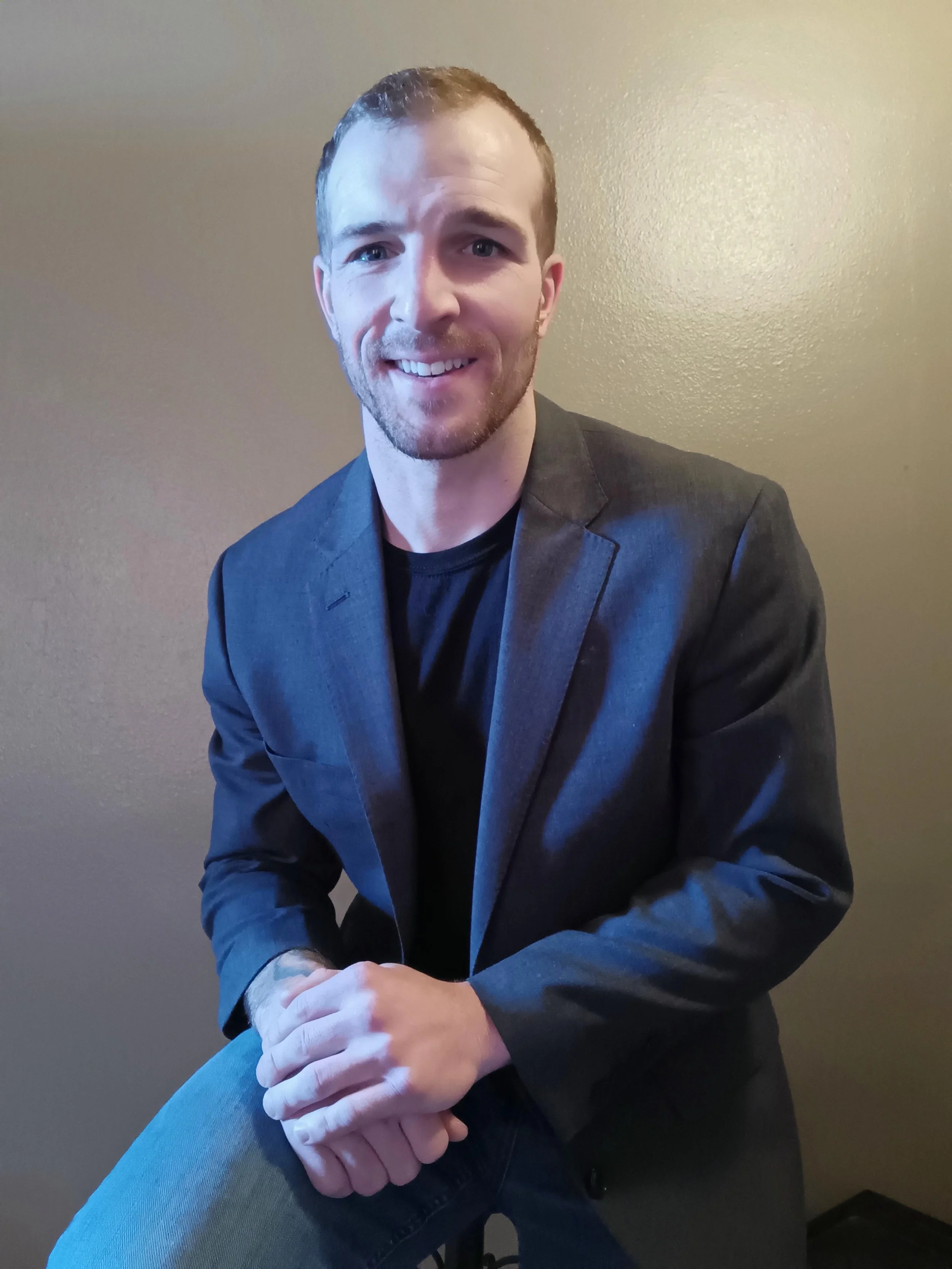 A young man with short brown hair, light skin, and a slight beard, smiling, wearing a dark blazer and black shirt, sitting against a beige wall.