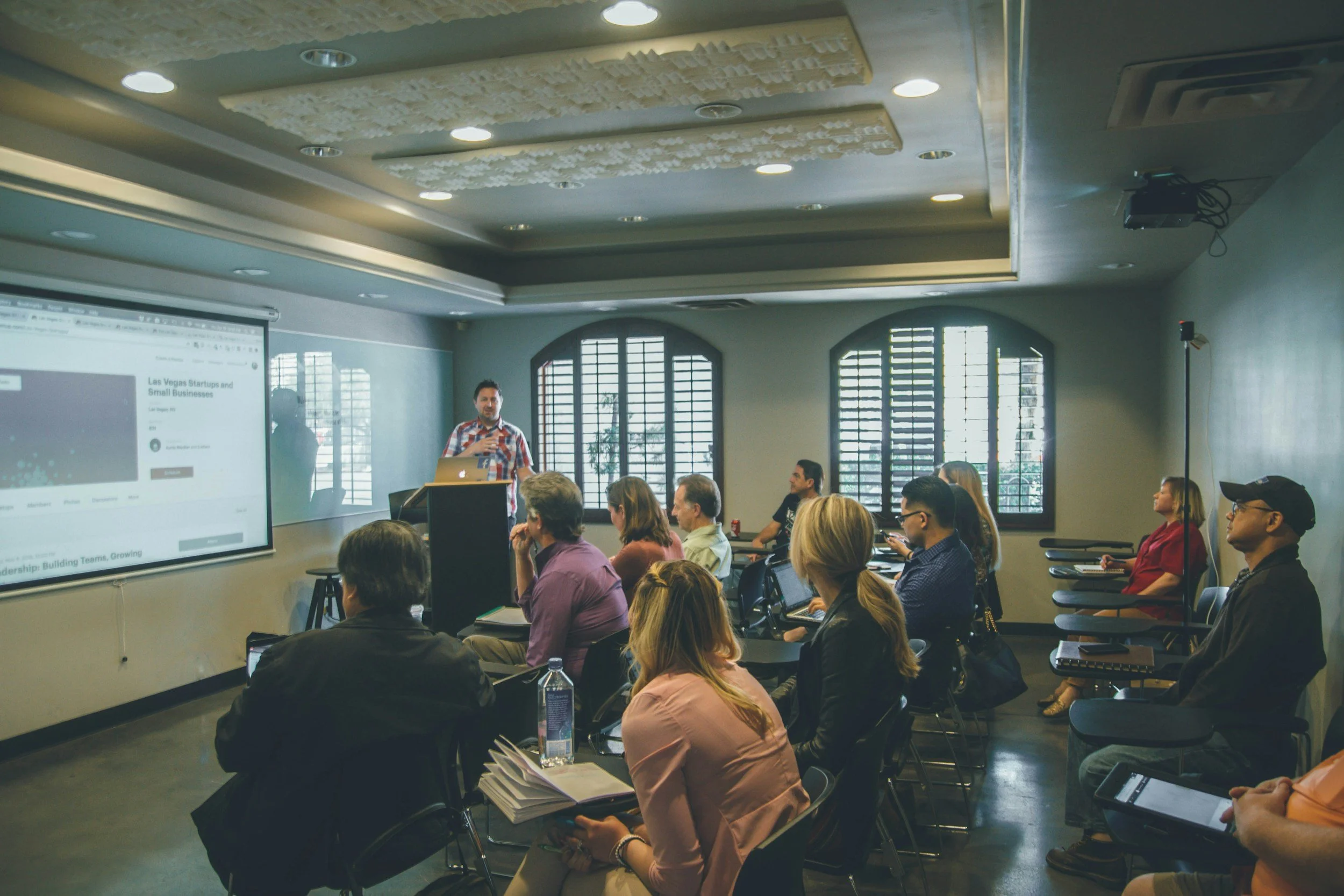 A man in a casual shirt giving a presentation to an audience in a conference room with arched windows and a large screen display.