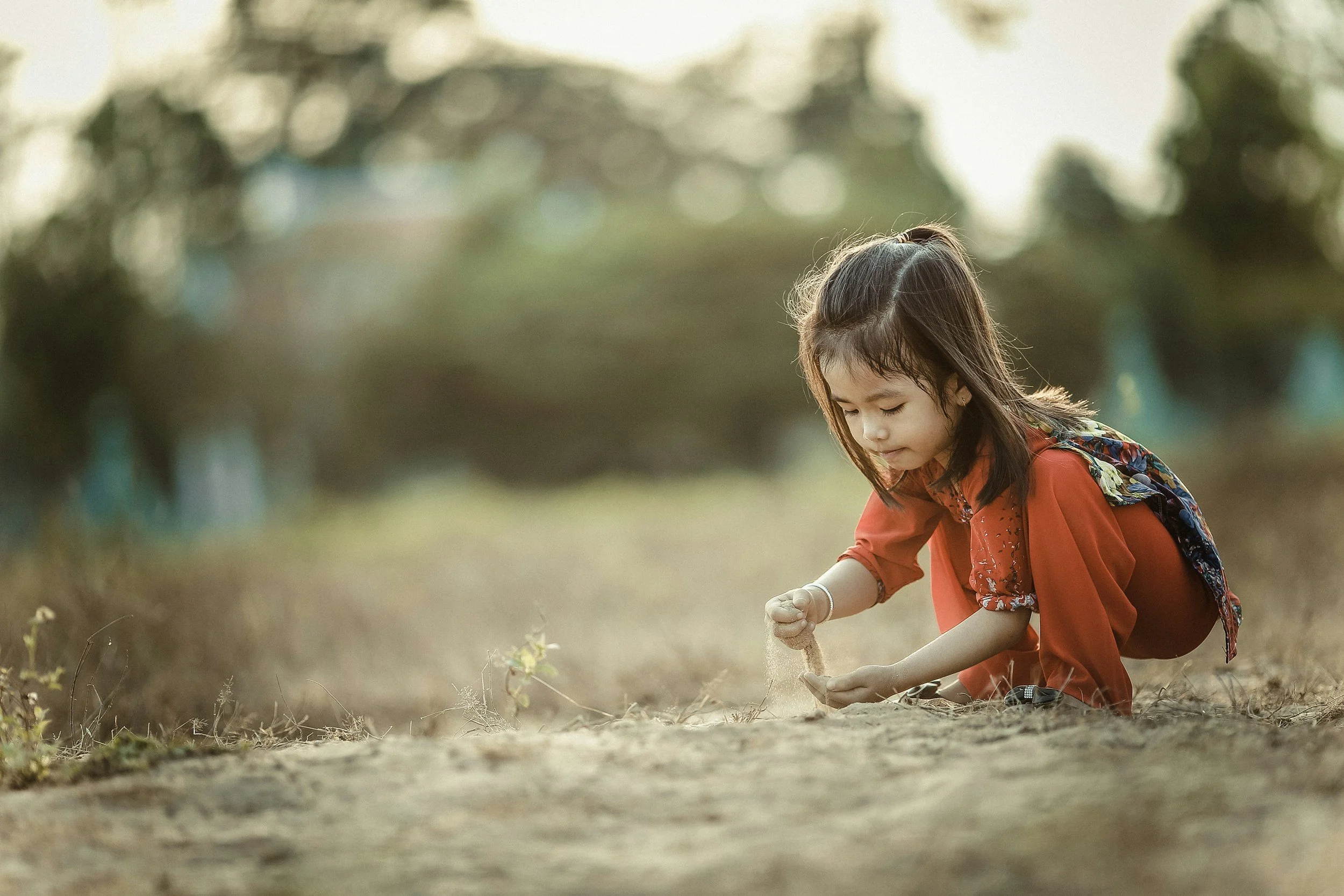 Young girl with dark hair in a ponytail, wearing a red coat, squatting and playing in the sand.