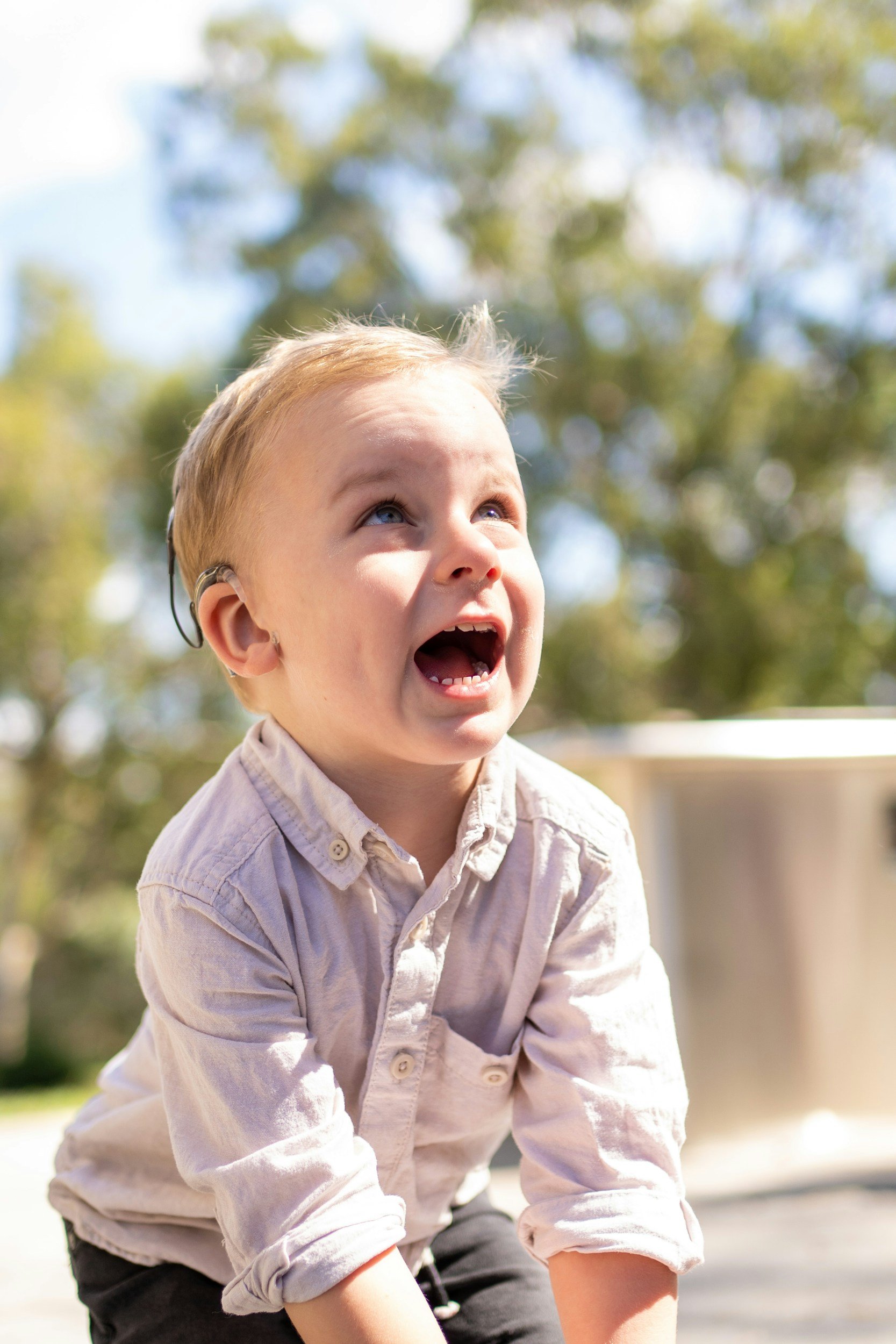 A young boy with blonde hair, wearing a beige button-up shirt and cochlear implants, looking upward with a joyful expression against a blurred outdoor background of trees and blue sky.
