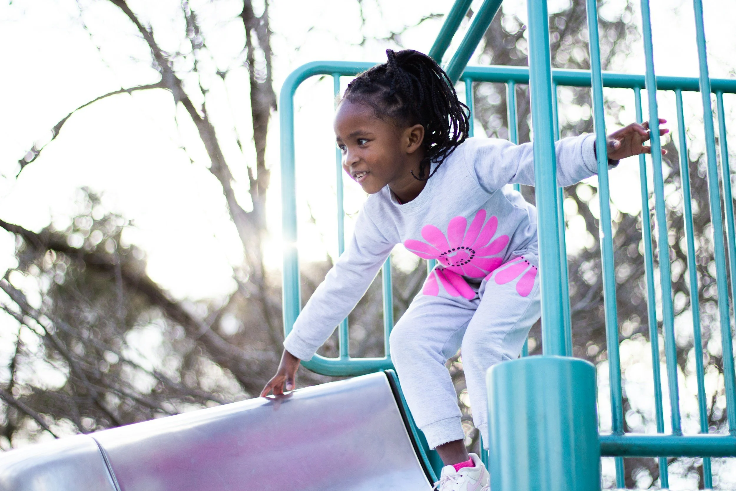A young girl with braided hair is climbing up a slide at a playground made of metal, surrounded by trees.