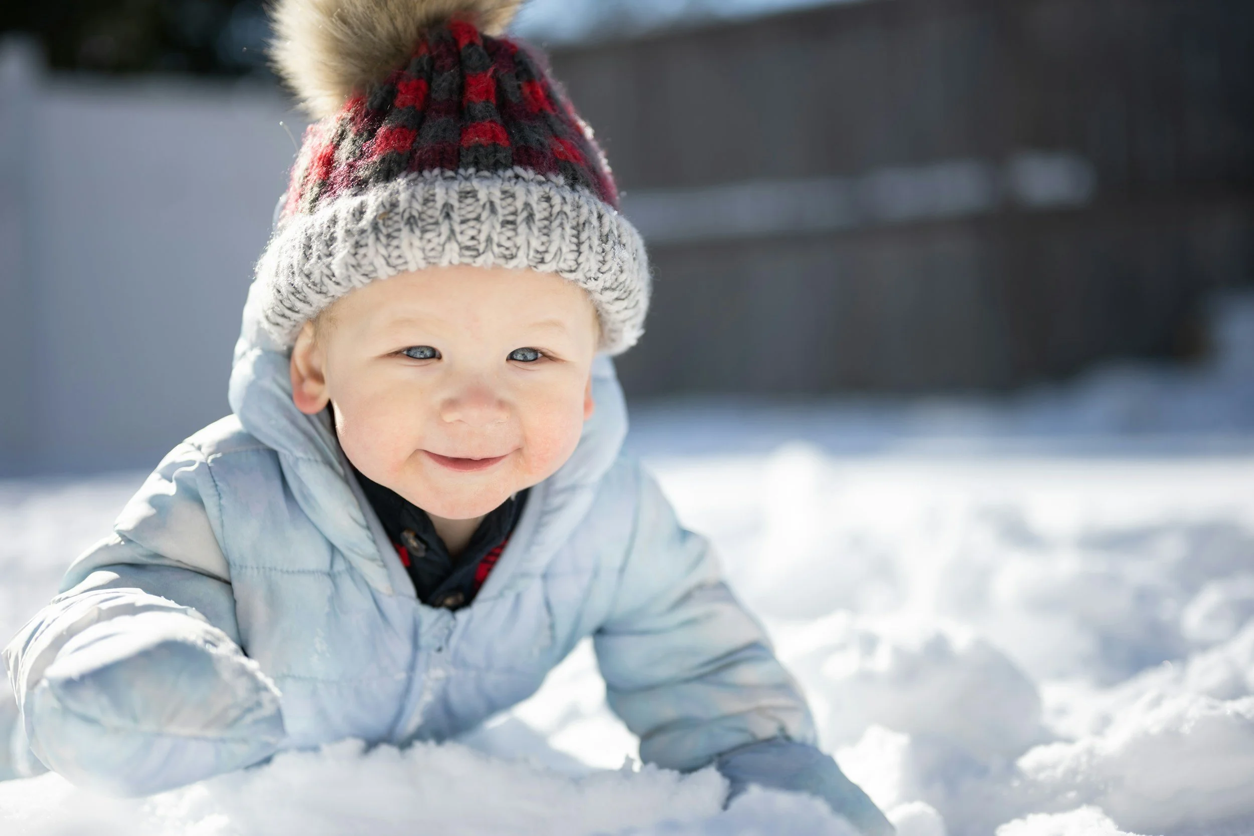 A young child with bright blue eyes and rosy cheeks playing in the snow, wearing a gray jacket and a red, black, and white knit hat with a pom-pom on top.