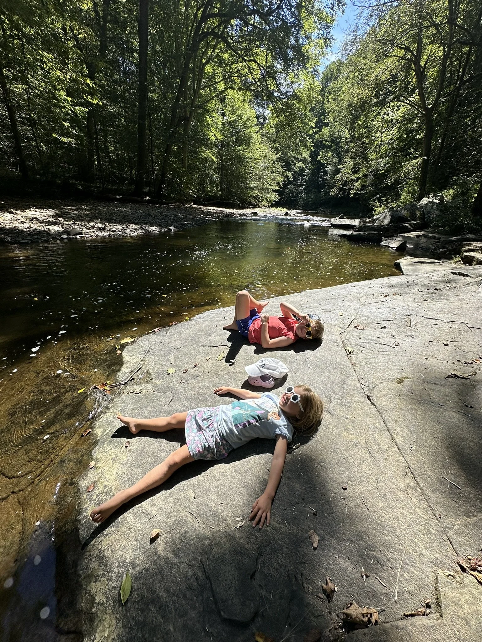Two children lying on a large rock by a river, surrounded by lush green trees, with both children wearing sunglasses, enjoying a sunny day outdoors.