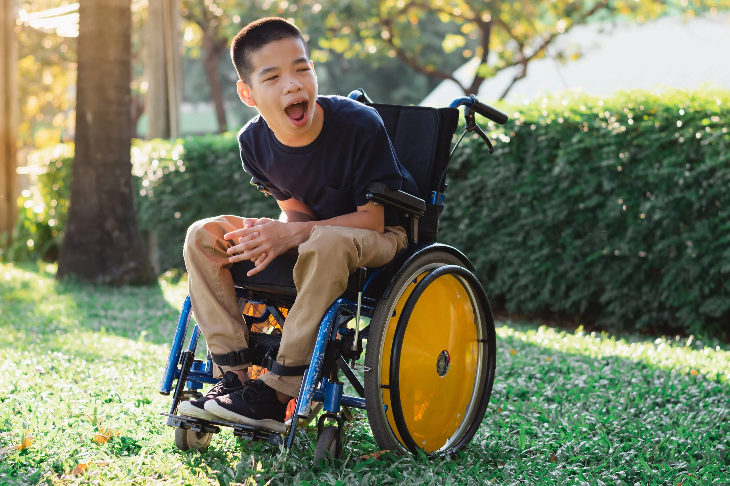 A young boy with short hair in a wheelchair outdoors on a grassy area, with an excited expression, wearing a dark T-shirt and tan pants. There are trees and bushes in the background on a sunny day.