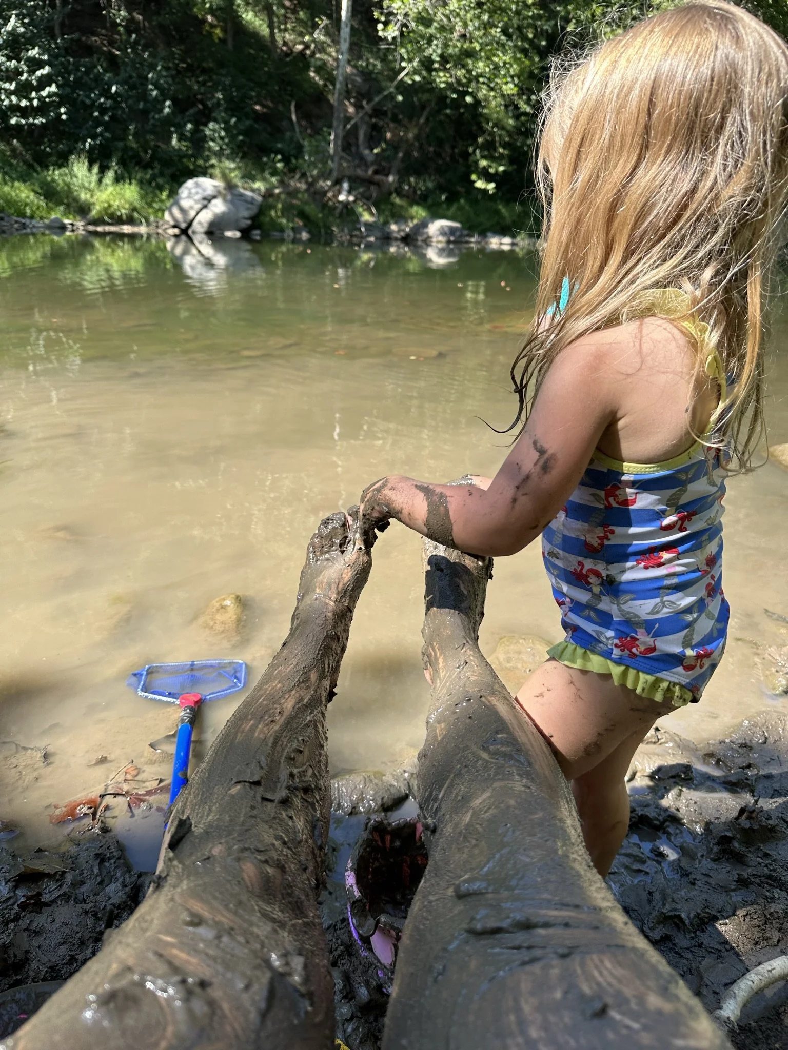 A young girl with blonde hair, dressed in a colorful swimsuit, sits on a muddy log in a shallow river or creek. She is playing with the mud, and a small blue fishing net lies nearby in the water. The background features green trees and rocks.