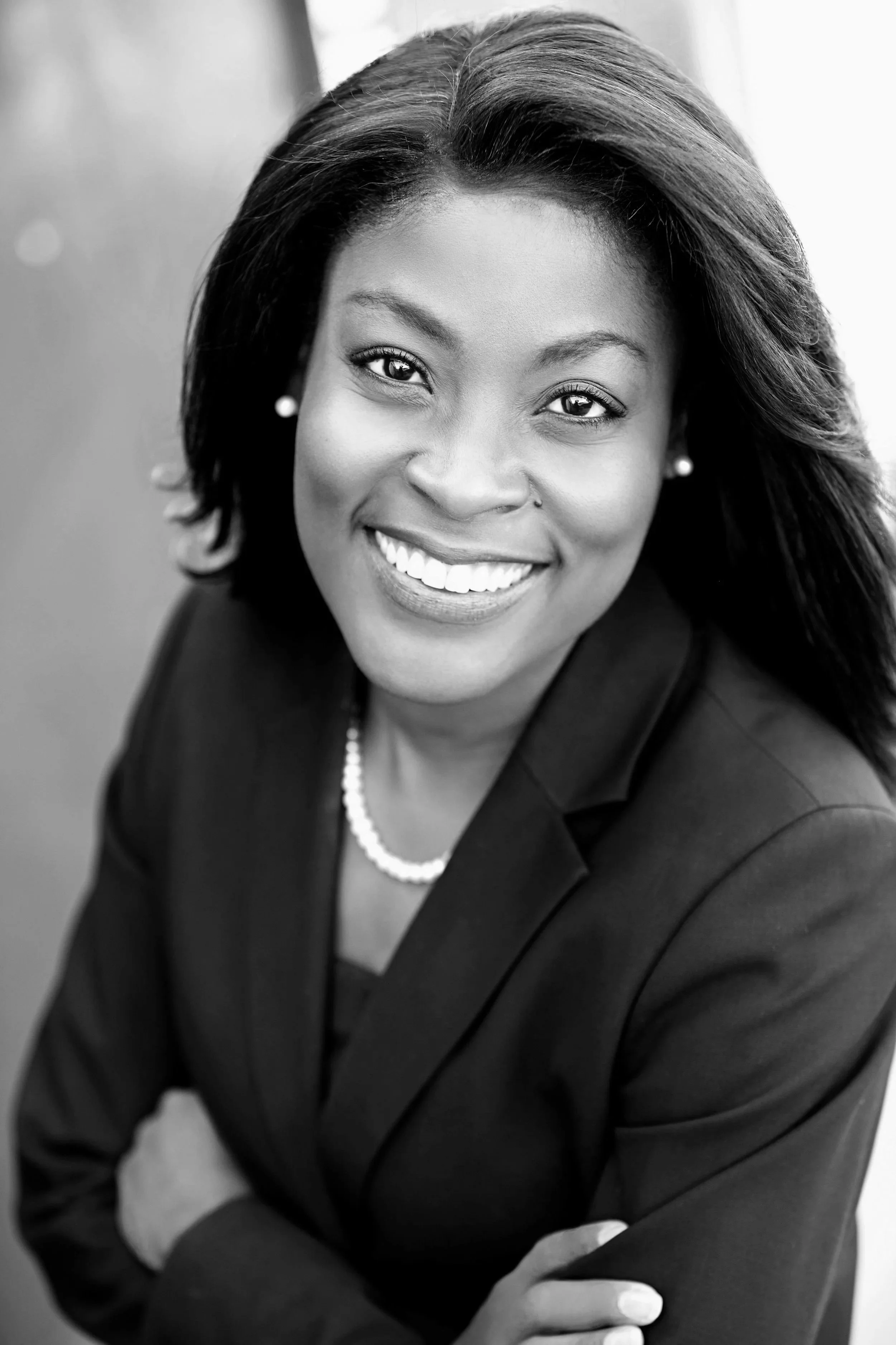 A black-and-white portrait of a woman with shoulder-length dark hair, smiling with visible teeth, wearing pearl earrings, a pearl necklace, and a dark blazer, with her arms crossed and a blurred background.