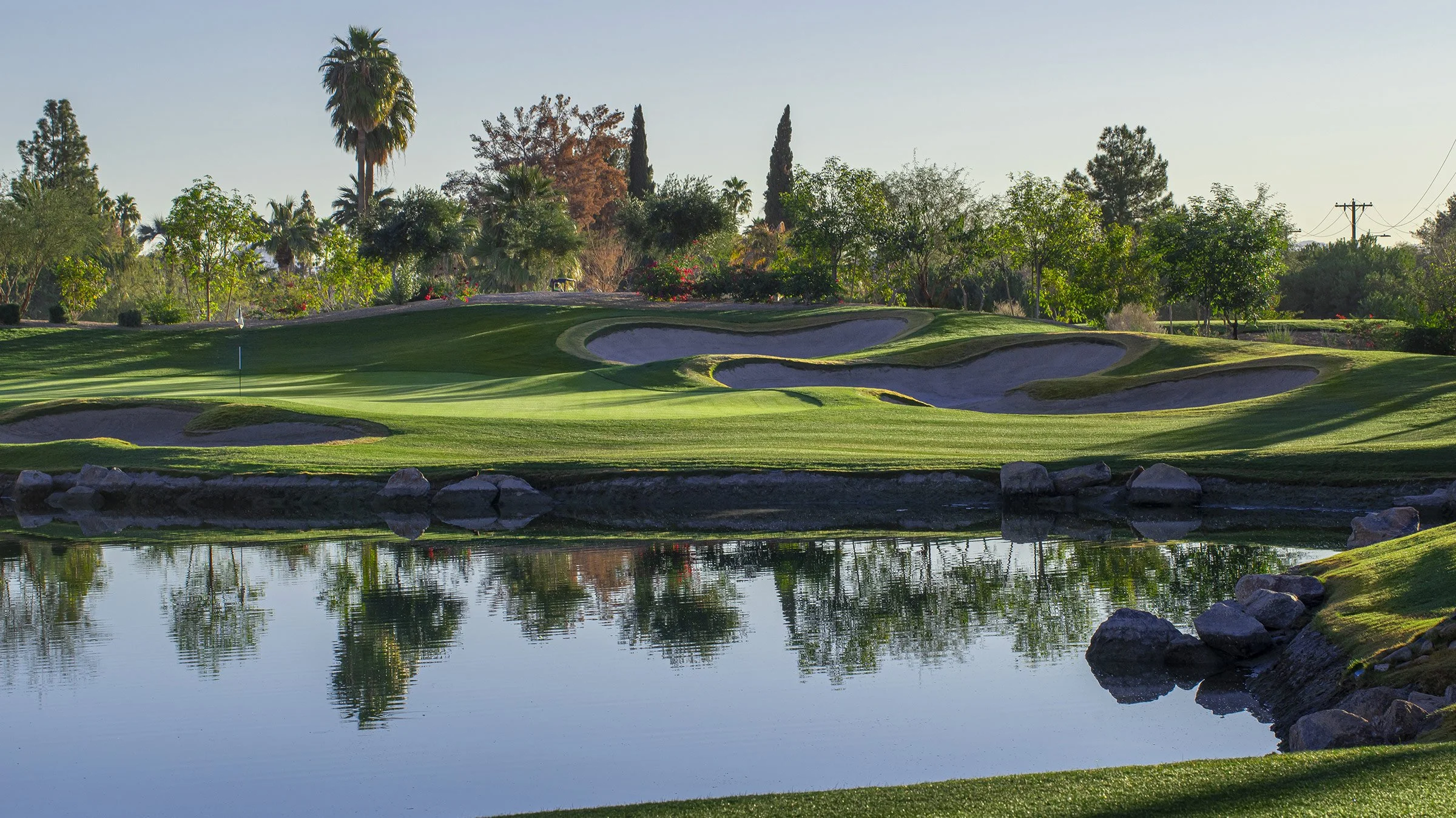A golf course with sand bunkers, green grass, and a water hazard, surrounded by trees and a clear sky.