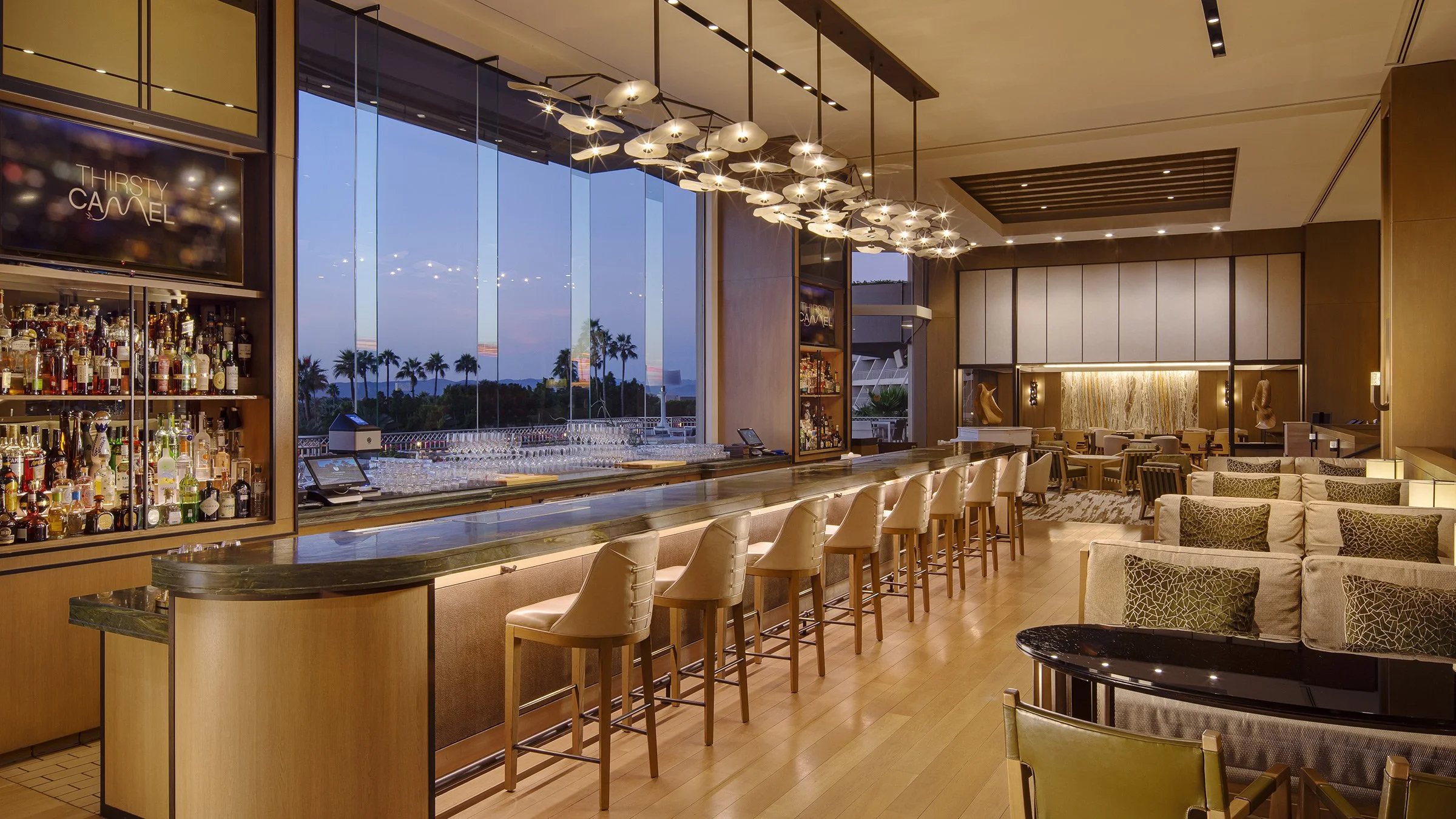 Interior of a modern bar and lounge area with a view of palm trees outside through large glass windows, featuring a sleek bar counter with chairs, a variety of bottles and glassware, contemporary lighting fixtures, and comfortable seating areas.