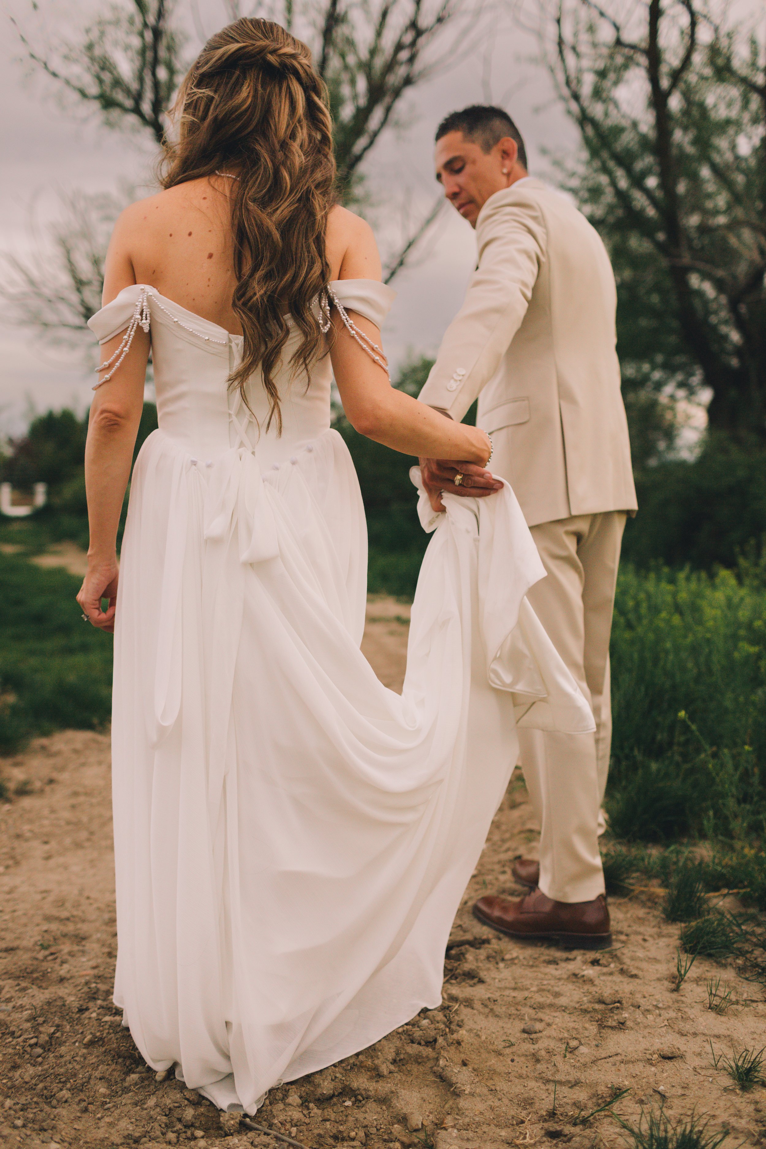 A bride and groom holding hands outdoors during her wedding, with the bride wearing a white off-shoulder dress with pearl embellishments and the groom in a cream suit, standing on a dirt path with trees in the background.