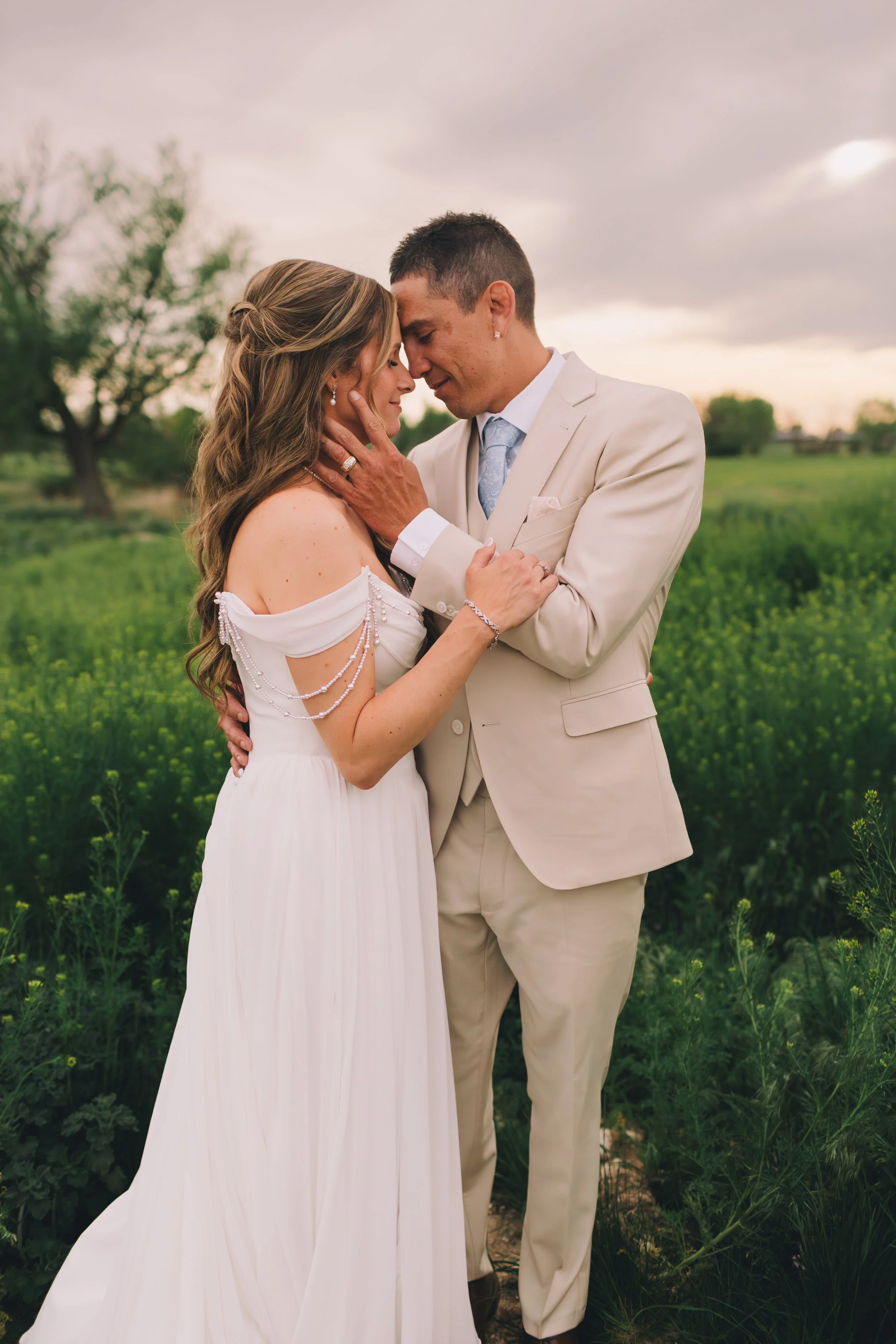 A bride and groom stand close together in a field, touching foreheads and holding each other tenderly during their wedding ceremony, with a cloudy sky and trees in the background.