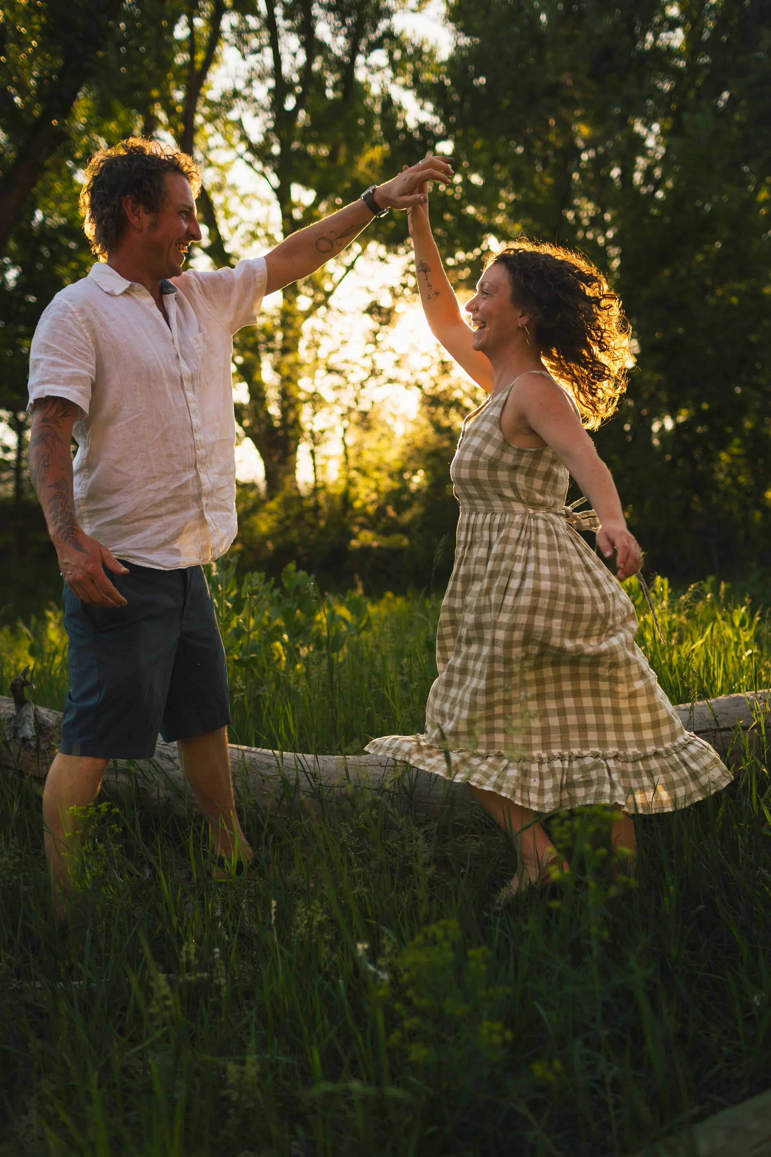 A smiling man and woman dancing outdoors at sunset, with trees in the background. The woman is wearing a checkered dress and the man is wearing a white short-sleeved shirt and dark shorts.